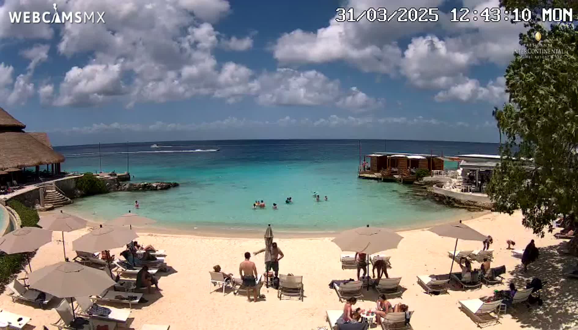 A sandy beach with turquoise water, several beach umbrellas and lounge chairs occupied by people, and a thatched-roof structure in the background under a partly cloudy sky.