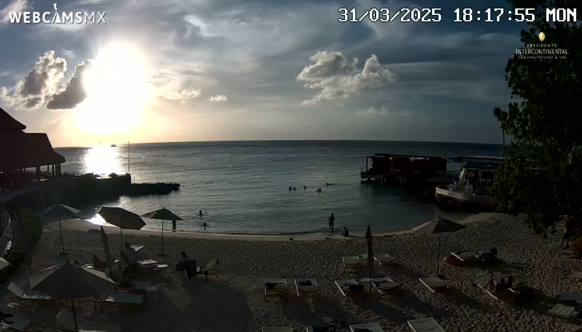 A beach scene at sunset shows a sandy beach with lounge chairs and umbrellas, calm ocean water with several people swimming, and a building on the shoreline under a partly cloudy sky.