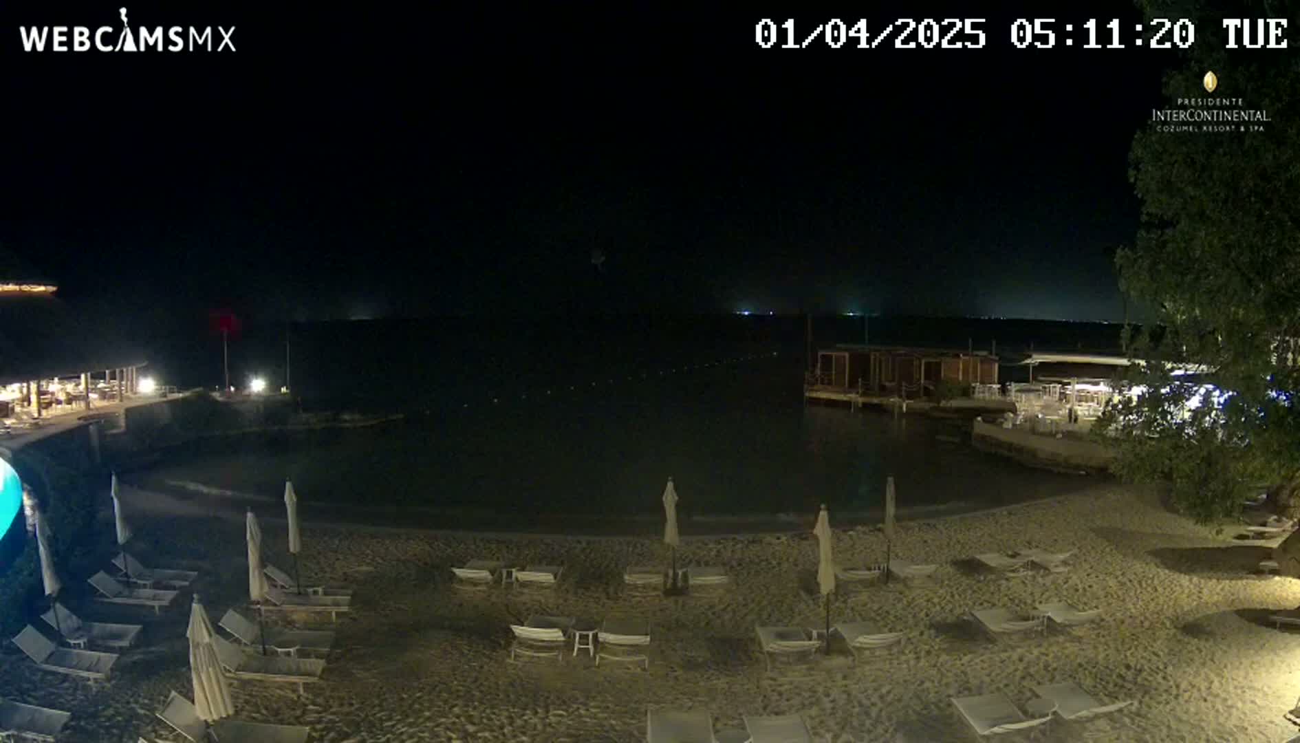 A nighttime beach scene shows calm water, empty lounge chairs under umbrellas on a sandy beach, and buildings with lights nearby.