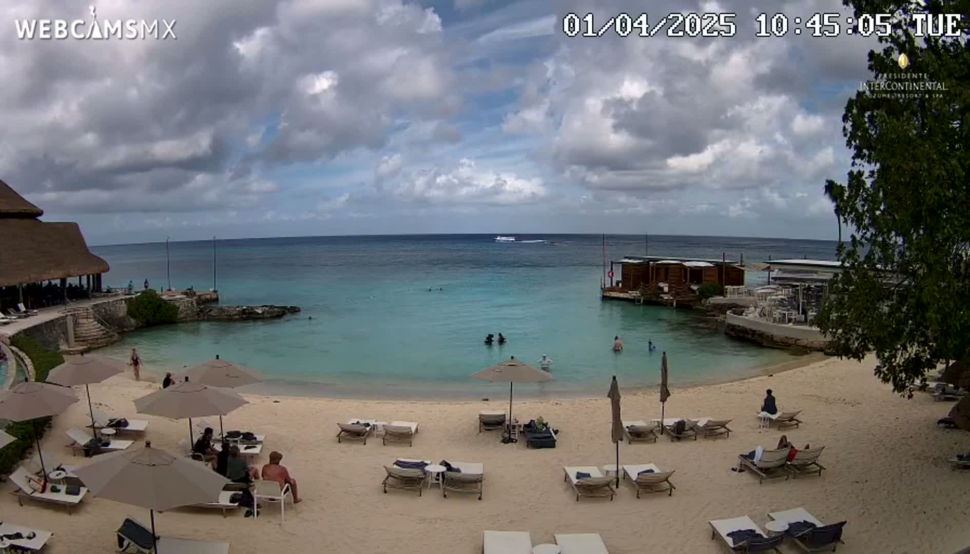 A partly cloudy day at a beach resort shows calm, turquoise water, a sandy beach with lounge chairs and umbrellas, and several people swimming and relaxing.