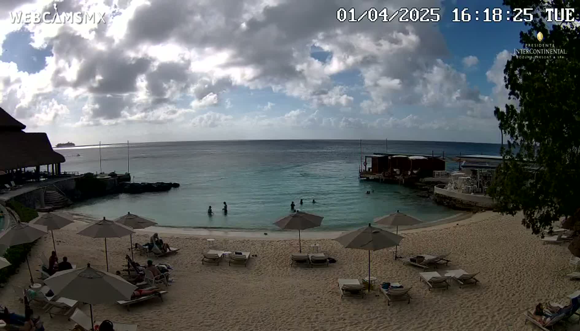 A partly cloudy beach scene shows calm, turquoise water with a few people swimming, beach chairs and umbrellas on a sandy beach, and buildings in the background.
