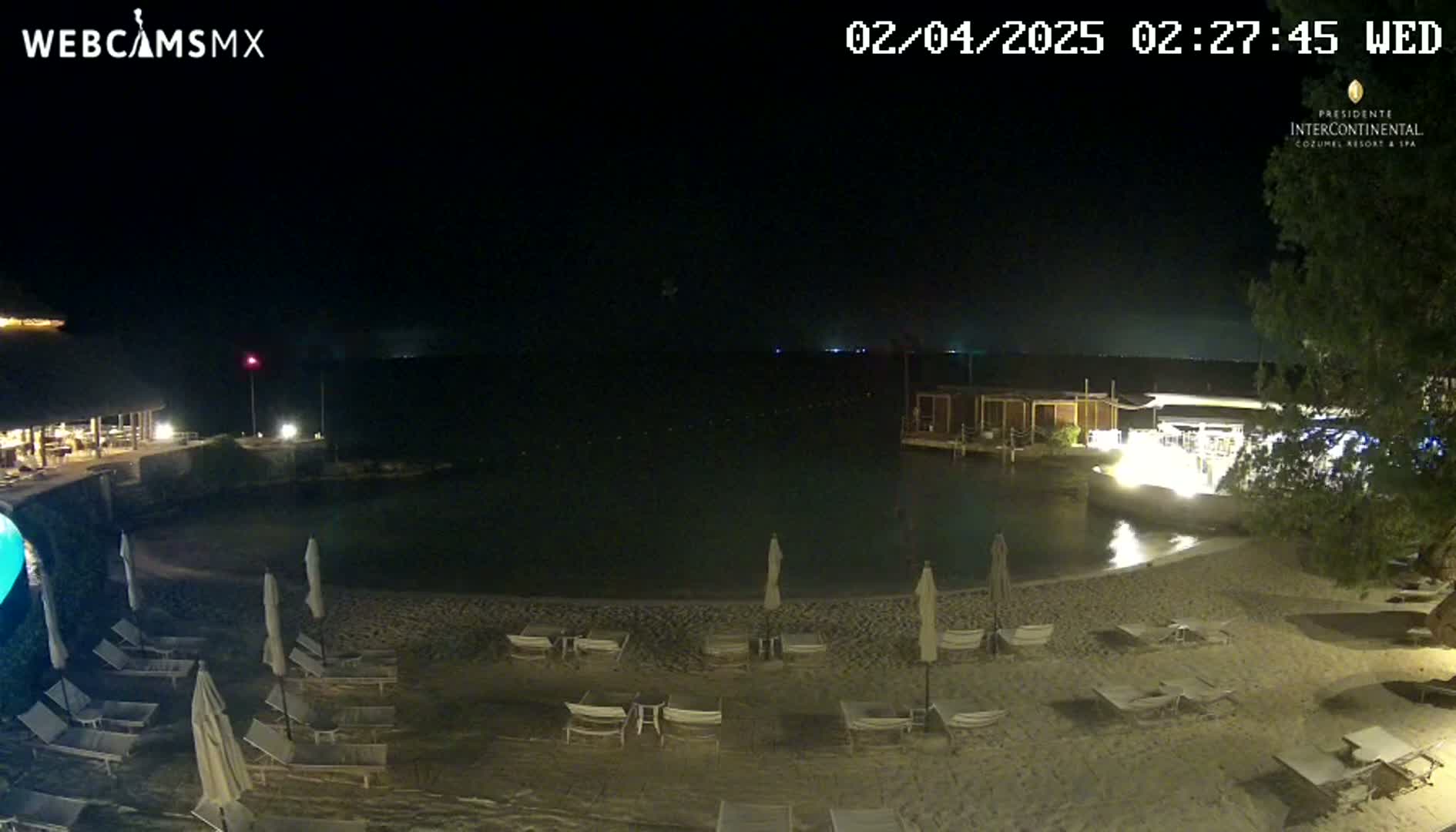 A nighttime beach scene shows a calm body of water, a sandy shoreline with empty lounge chairs and umbrellas under artificial lighting, and buildings in the background.
