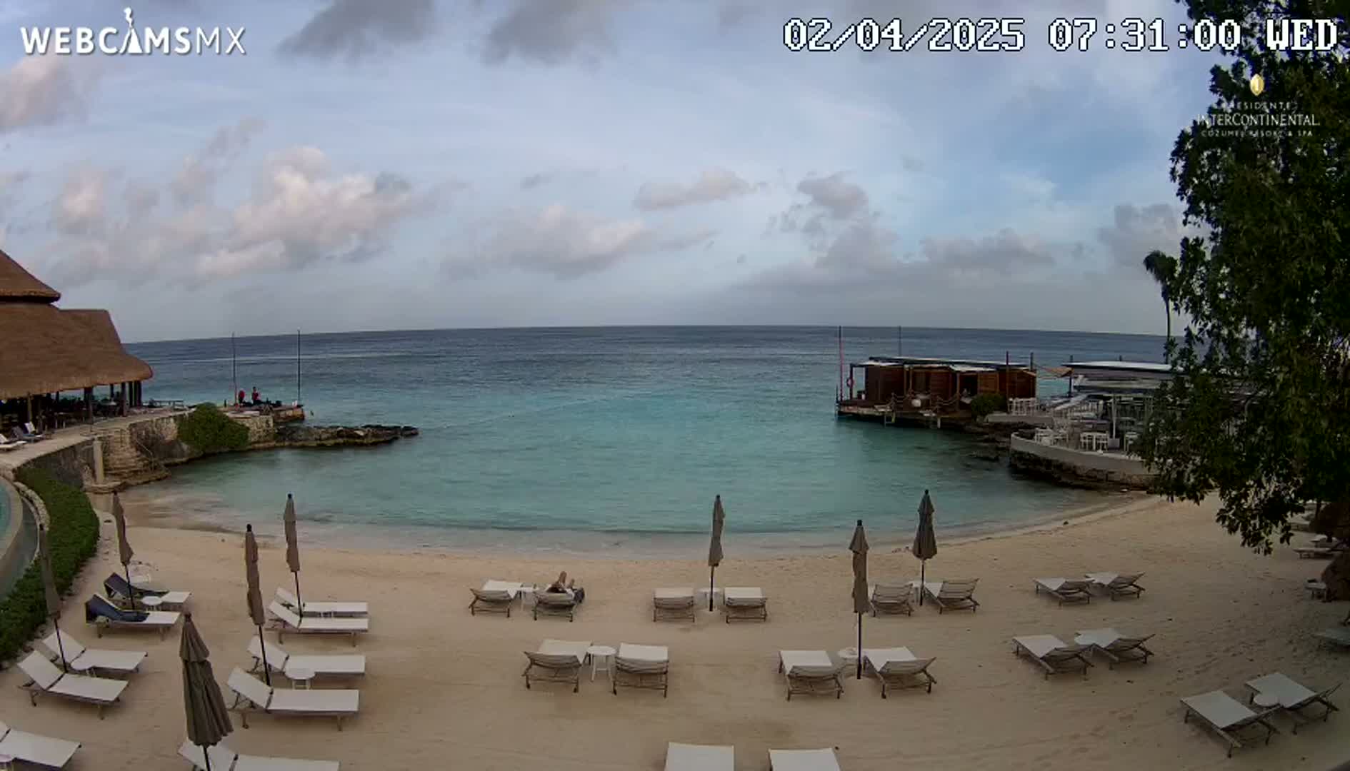 A partly cloudy day reveals a tranquil beach scene with lounge chairs, umbrellas, and a calm turquoise ocean, featuring a small overwater structure in the distance.