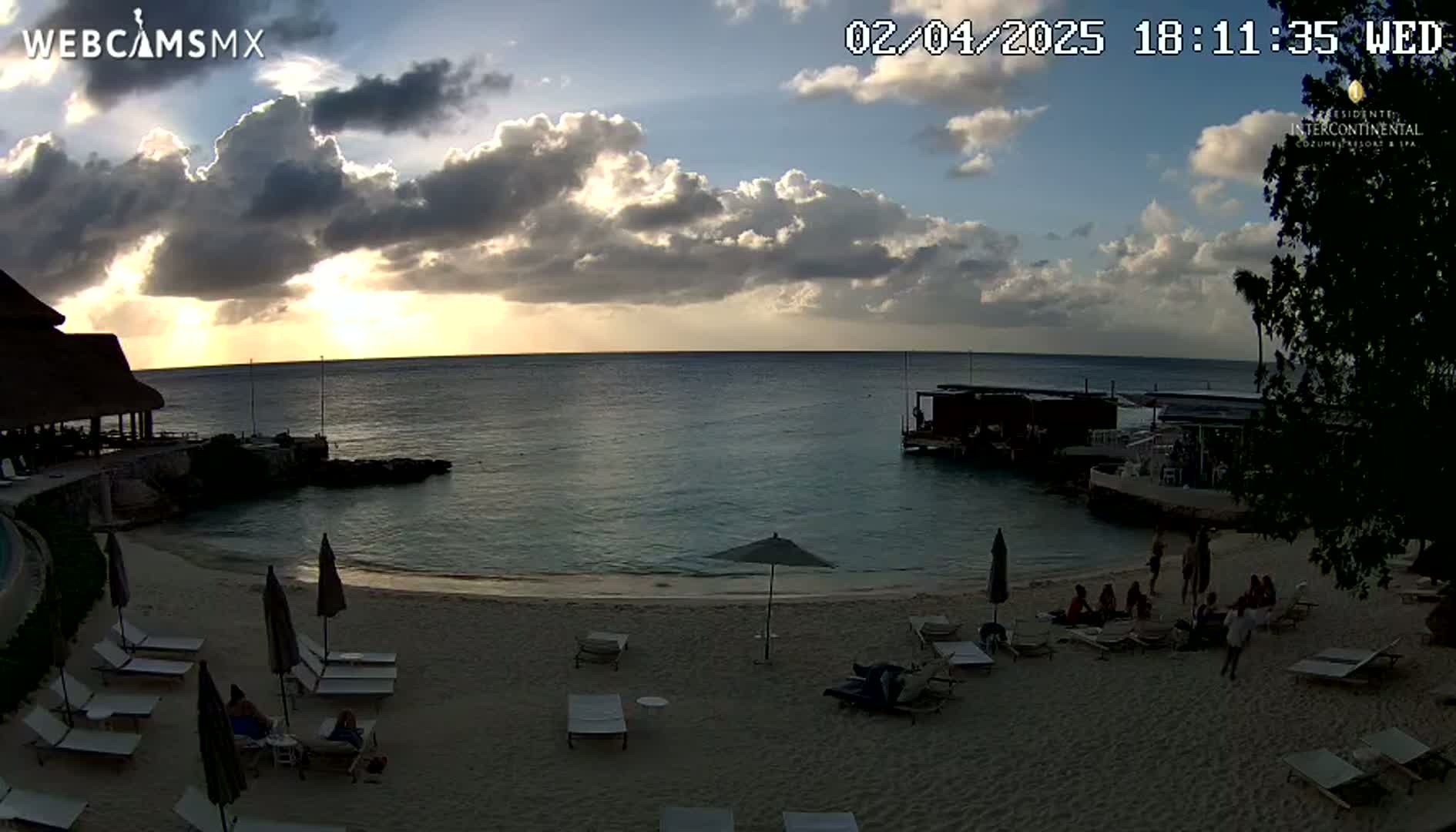 A beach scene at sunset, with partly cloudy skies, shows people relaxing on lounge chairs near the water's edge.