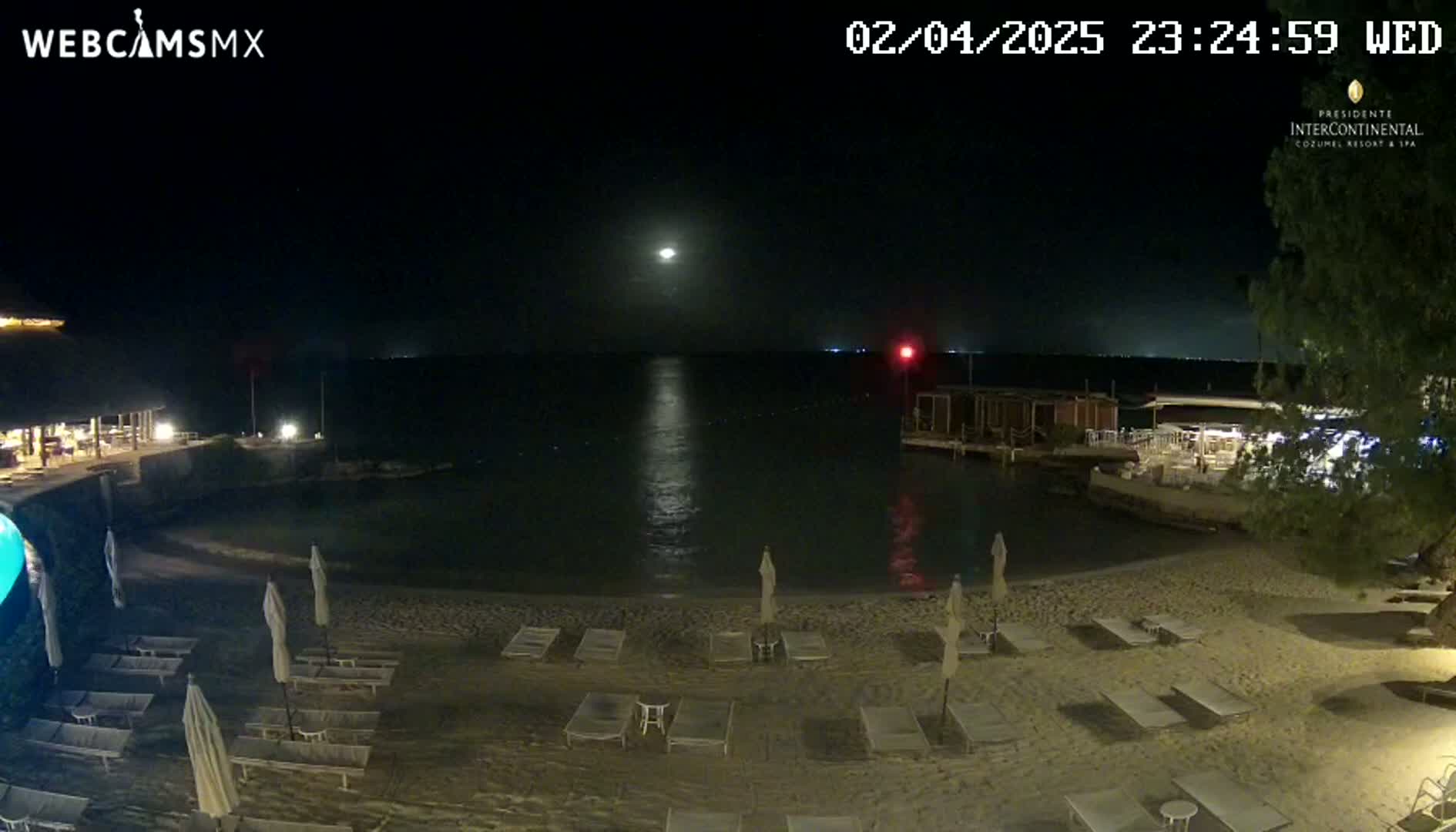 A moonlit beach at night, with lounge chairs and umbrellas arranged on the sand, overlooks calm water and a distant building.
