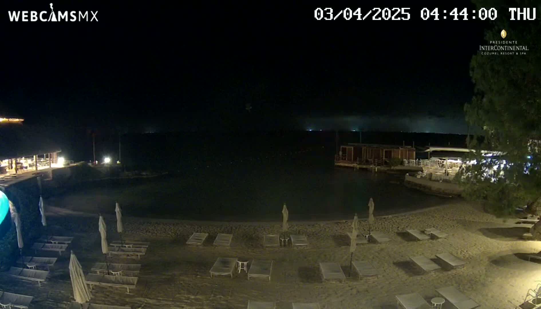 A nighttime beach scene shows a sandy shore with lounge chairs and umbrellas under a dark sky with some distant lights on the water.