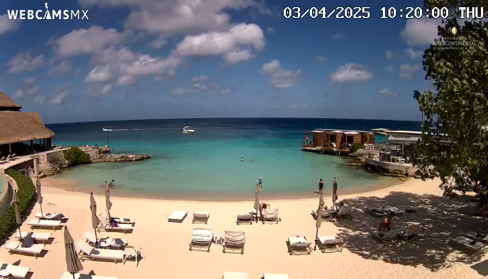 A sandy beach with lounge chairs and people enjoying a sunny day with partly cloudy skies, calm turquoise water, and a small boat in the distance.