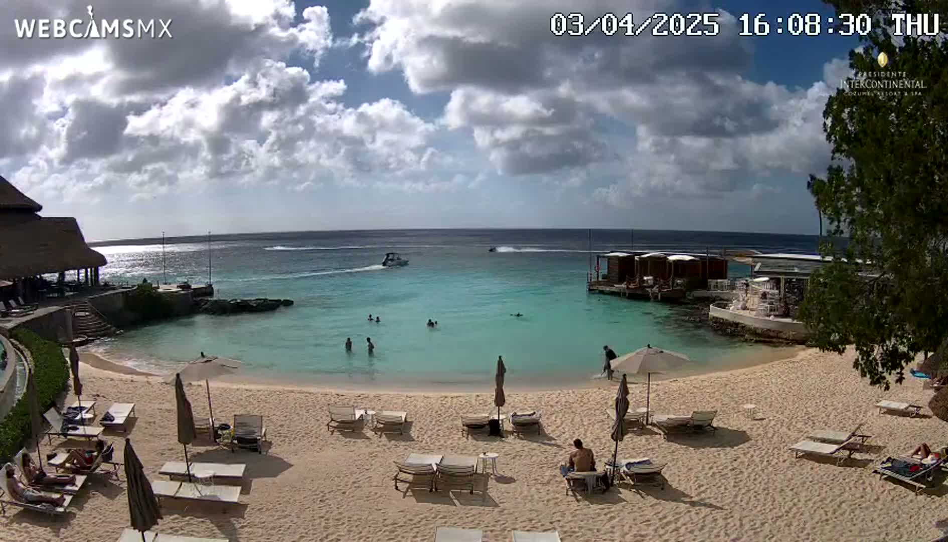 A partly cloudy sunny day shows a tranquil beach scene with calm turquoise water, a sandy shore lined with lounge chairs and umbrellas, and several people swimming and relaxing.