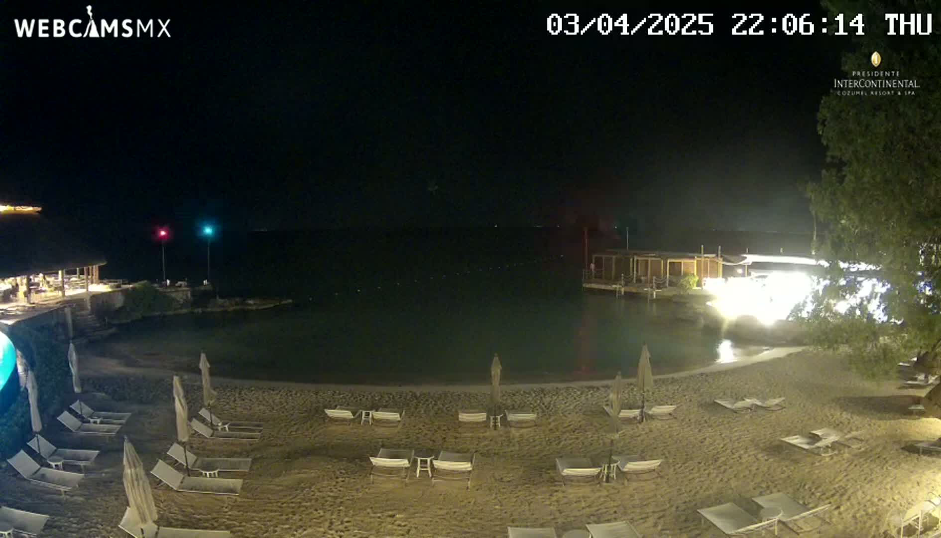 A nighttime beach scene shows a calm body of water next to a sandy shore with lounge chairs and umbrellas under a clear, dark sky.