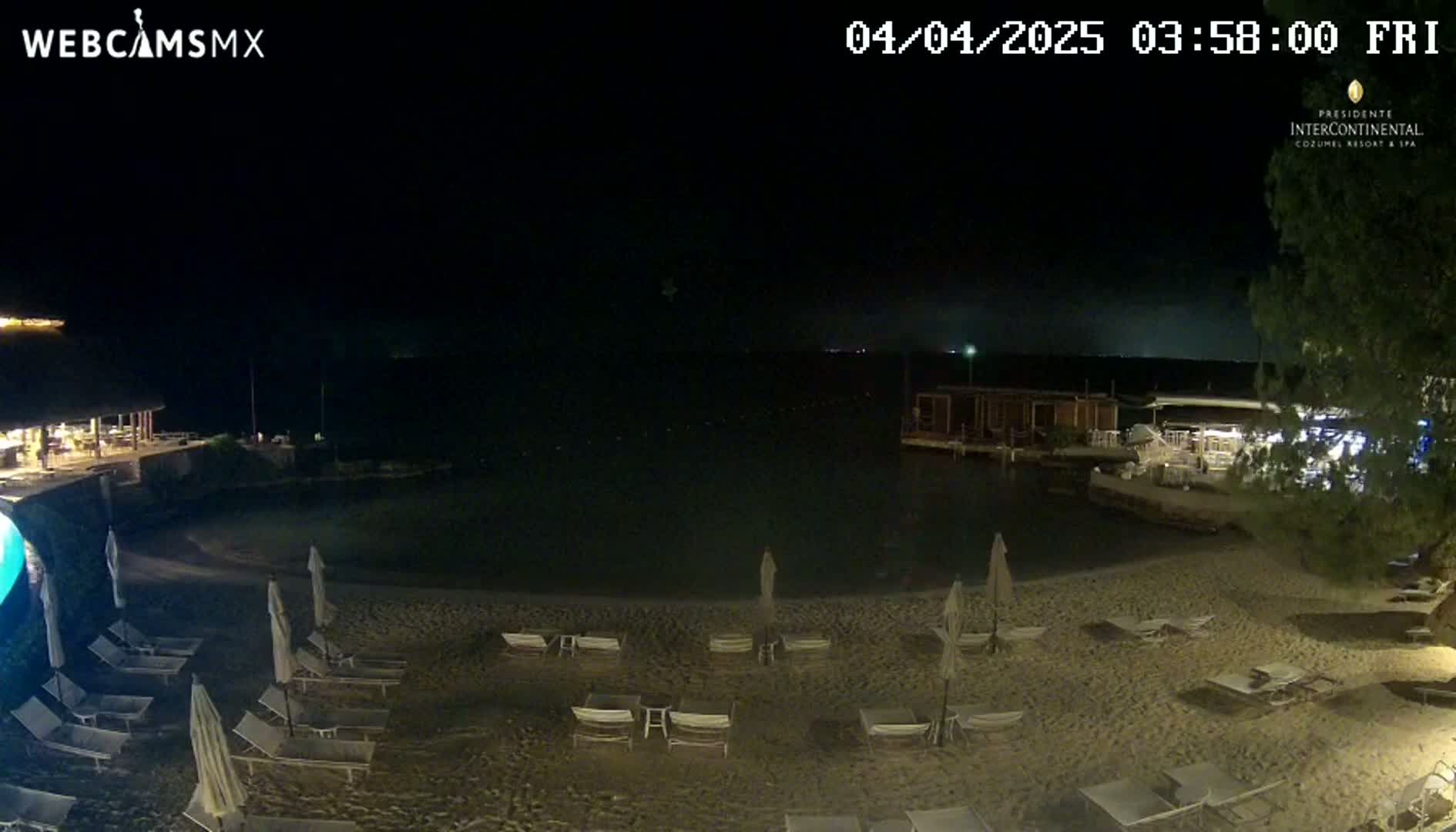 A nighttime beach scene shows a sandy shoreline with lounge chairs and umbrellas under a clear, dark sky, with calm water and buildings visible in the background.