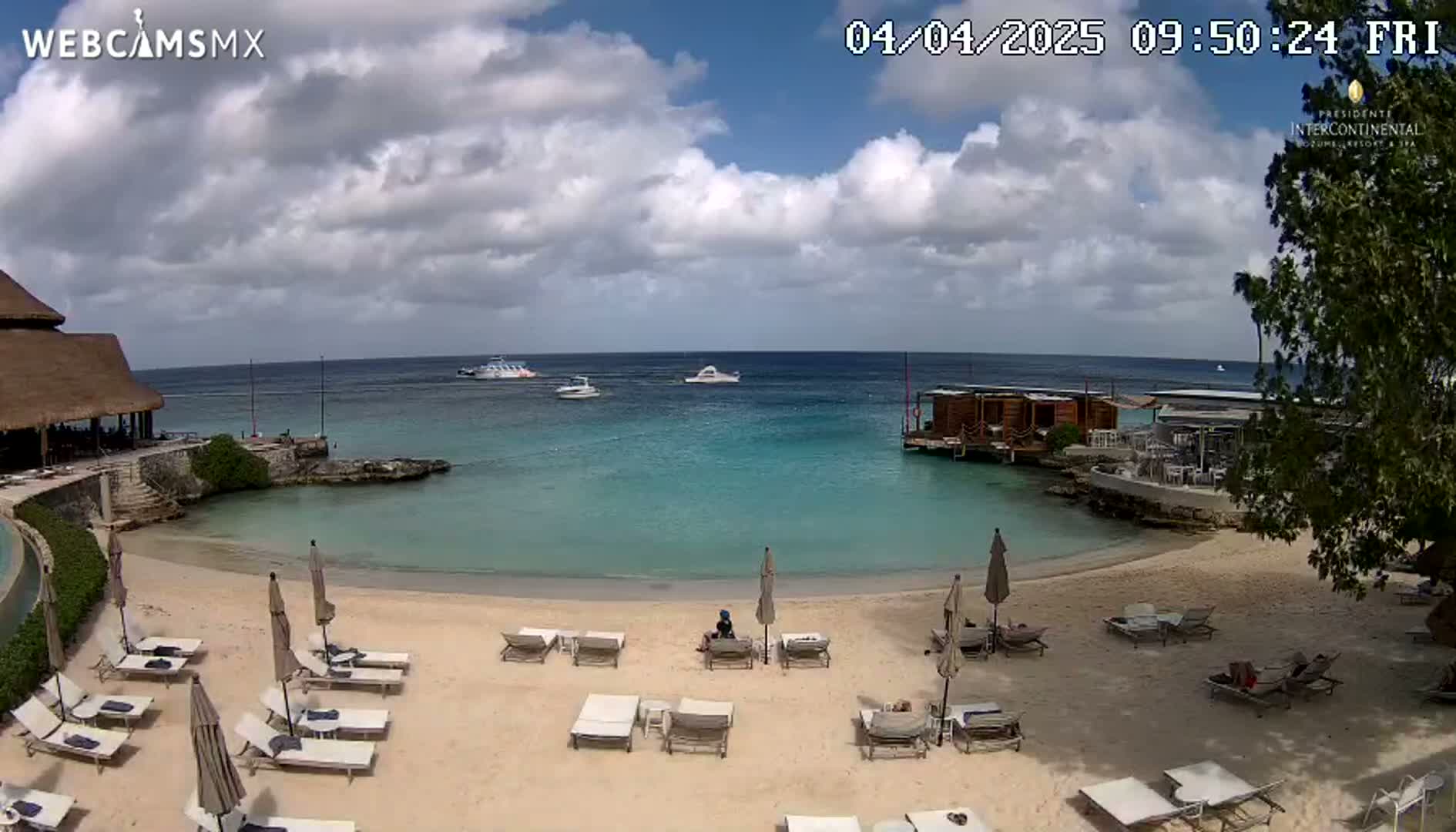 A partly cloudy day at a beach resort shows a sandy beach with lounge chairs and umbrellas, calm turquoise water, and several boats in the distance.