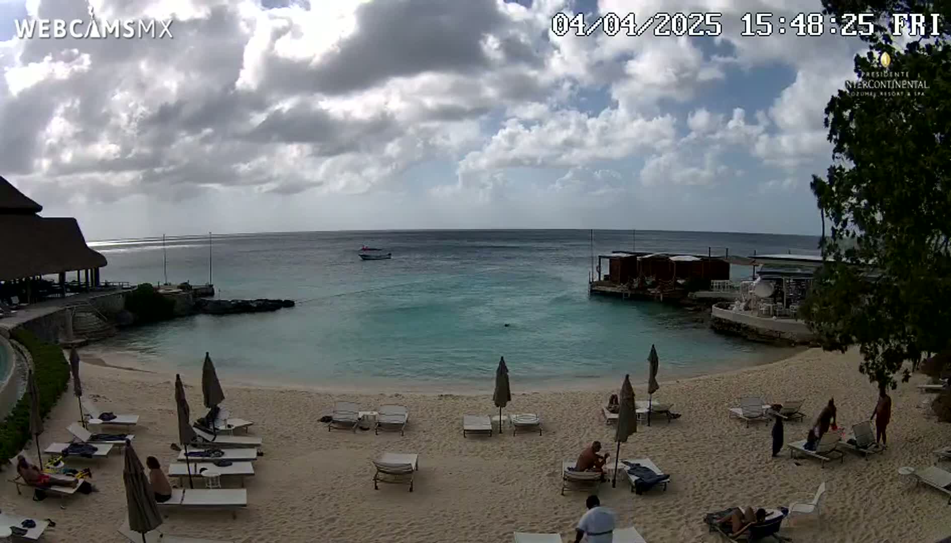 A partly cloudy day at a beach with calm, turquoise water, people relaxing on lounge chairs under umbrellas, and a few small boats visible in the distance.