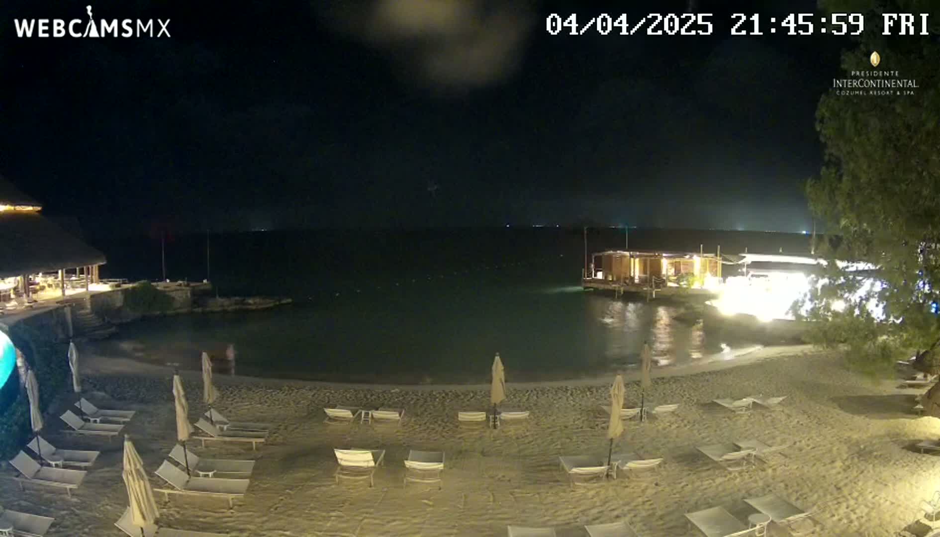 A nighttime beach scene shows empty lounge chairs on a sandy shore next to a calm body of water under a partly cloudy sky.