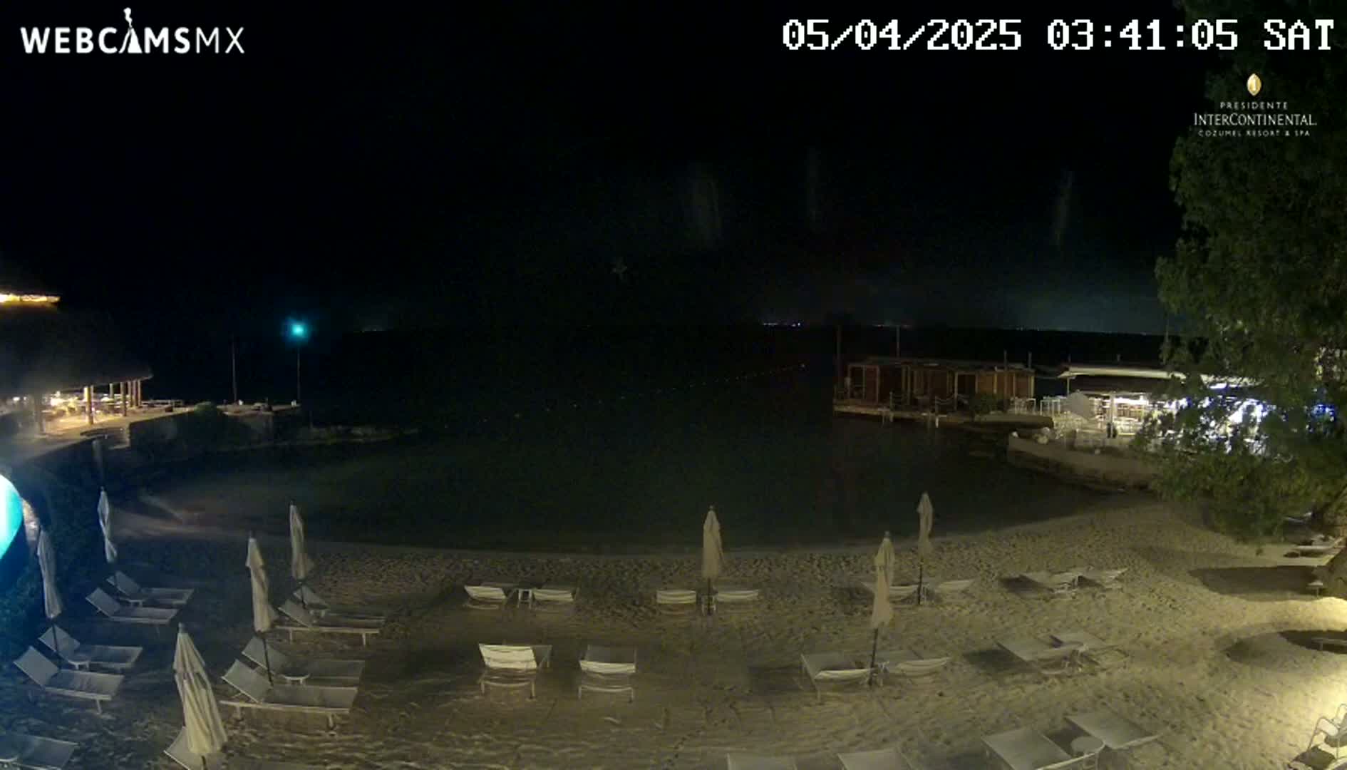 A nighttime beach scene shows several empty lounge chairs and umbrellas on a sandy beach next to calm water, with buildings and vegetation visible in the background under a clear, dark sky.
