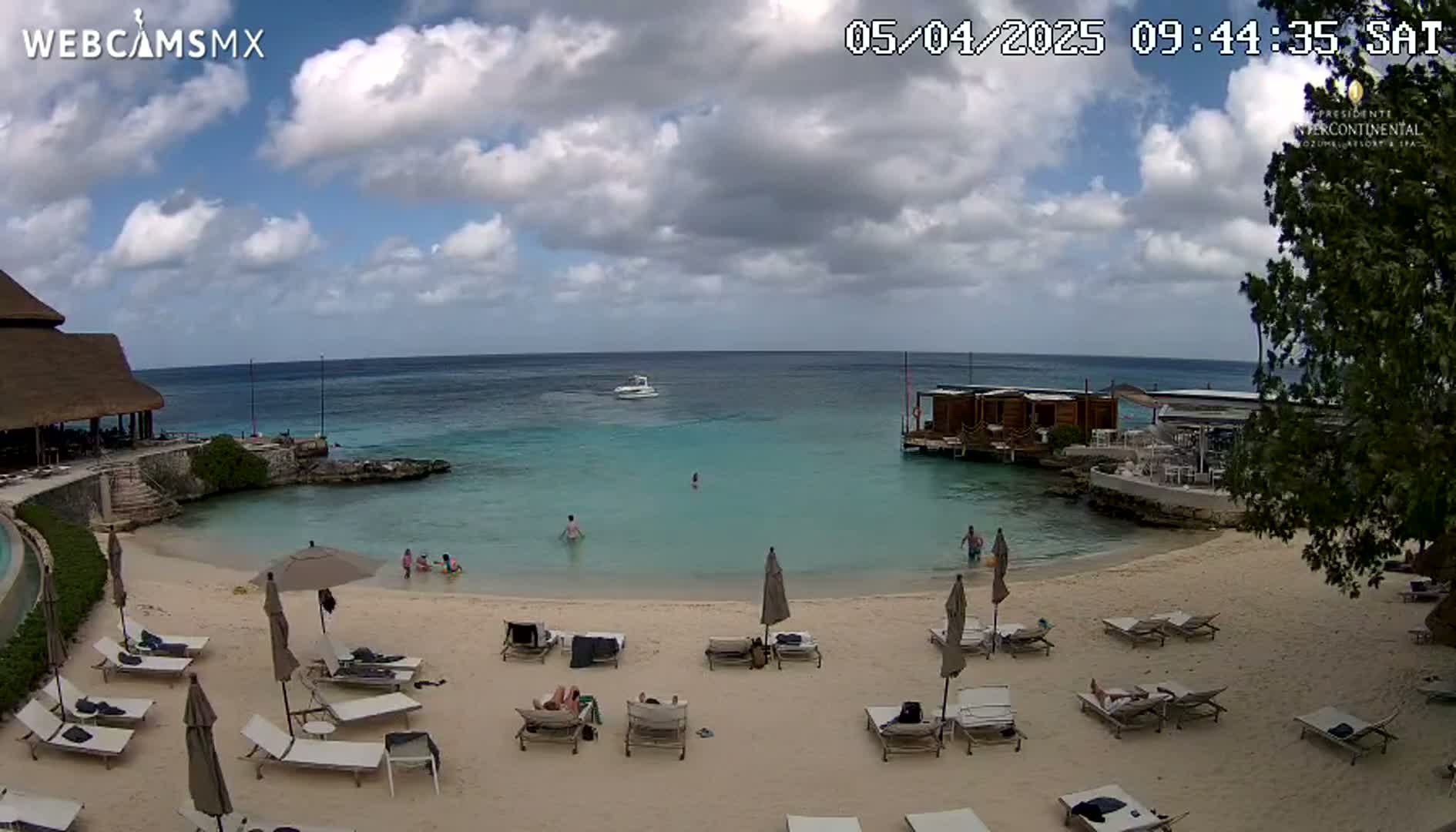 A partly cloudy day at a beach resort with calm turquoise water, a sandy beach with lounge chairs and umbrellas, and a few people swimming and relaxing.