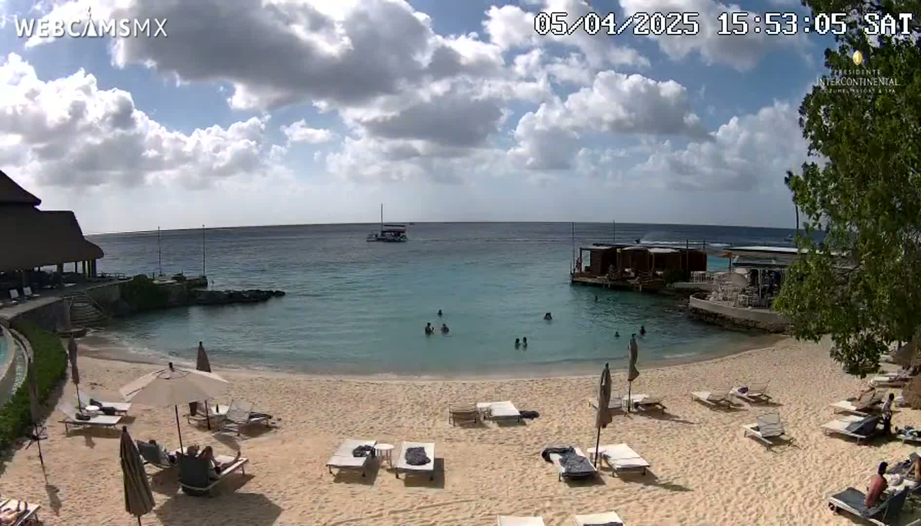 A partly cloudy day at a beach with calm, clear water, people swimming and sunbathing on lounge chairs under umbrellas.