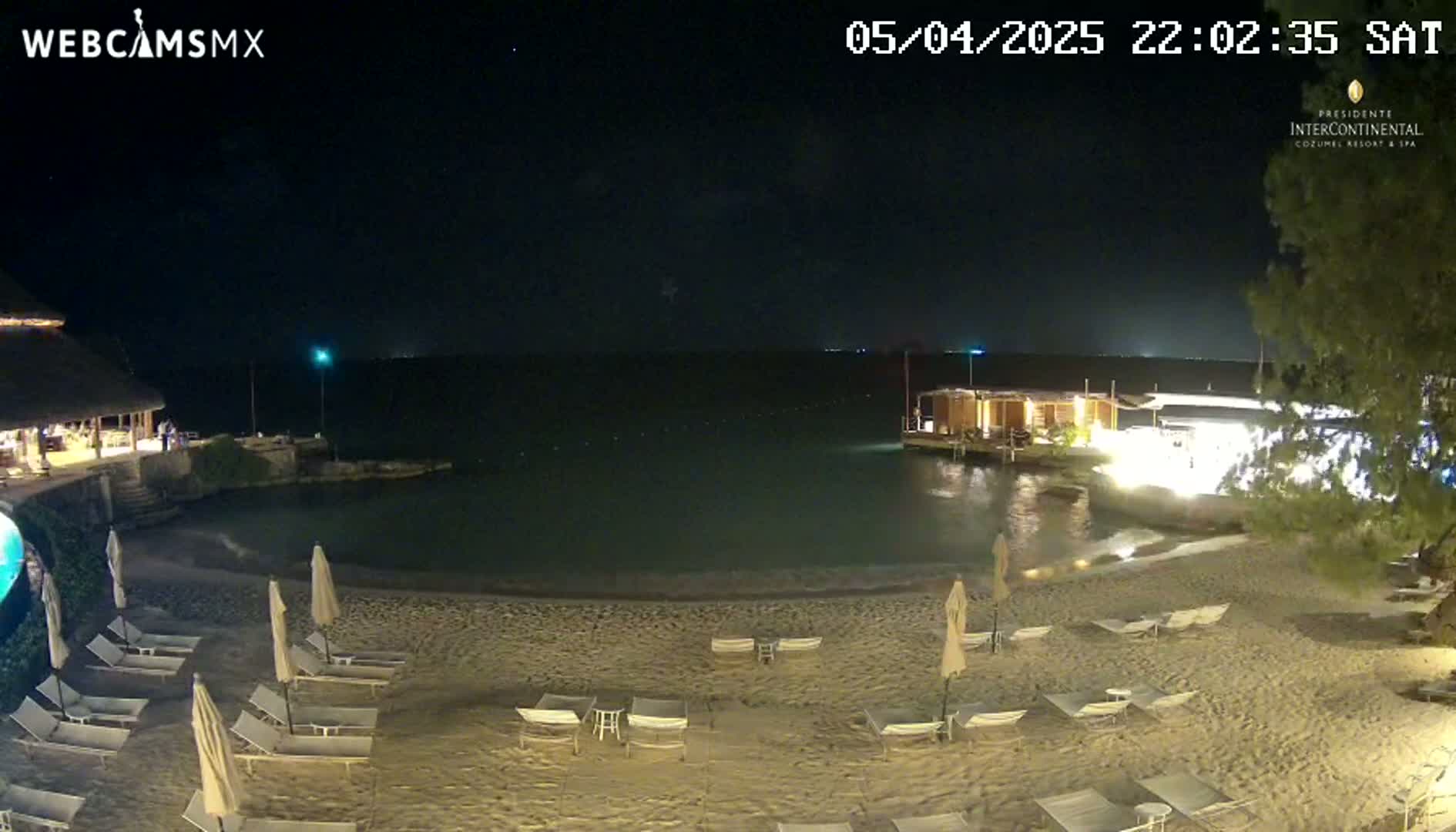 A nighttime beach scene shows a sandy shore with empty lounge chairs and umbrellas under a clear, dark sky.