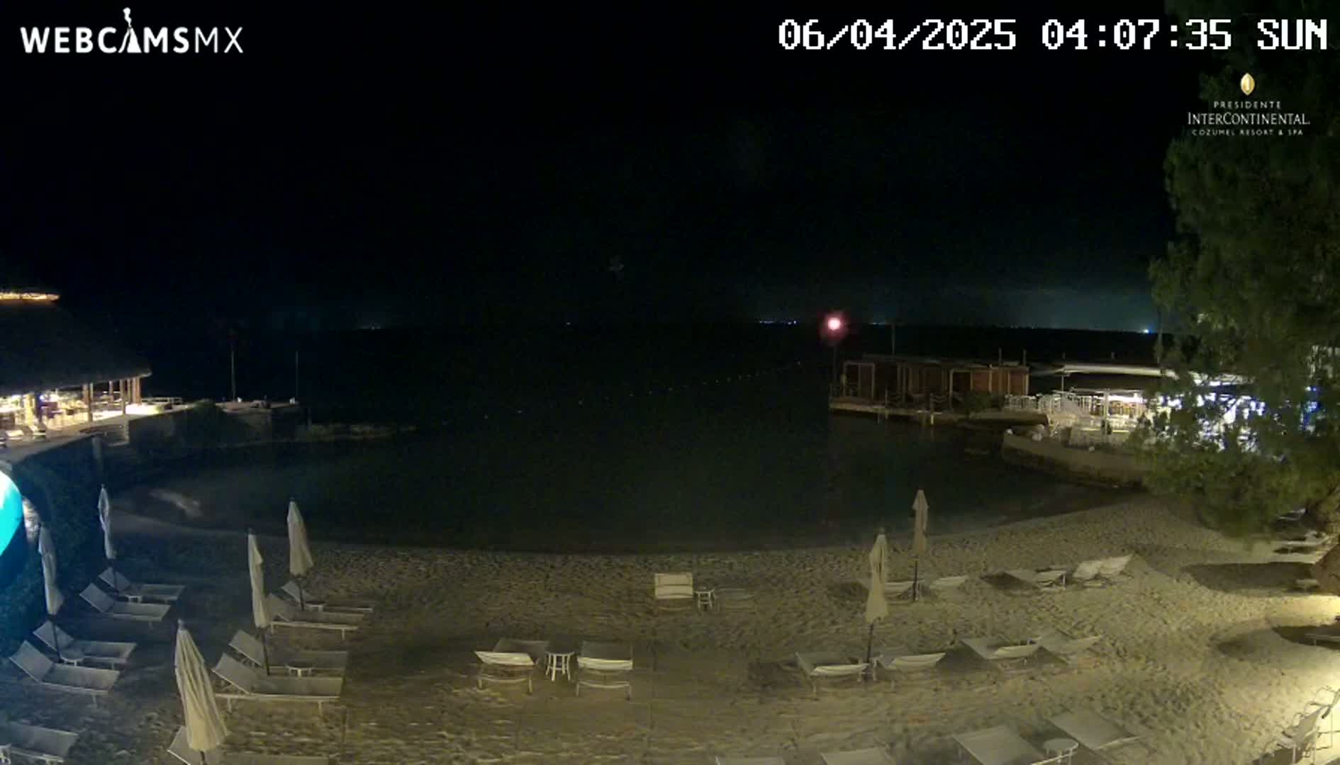 A nighttime view of a sandy beach with lounge chairs and umbrellas, a calm body of water, and low-lying buildings under a clear, dark sky.