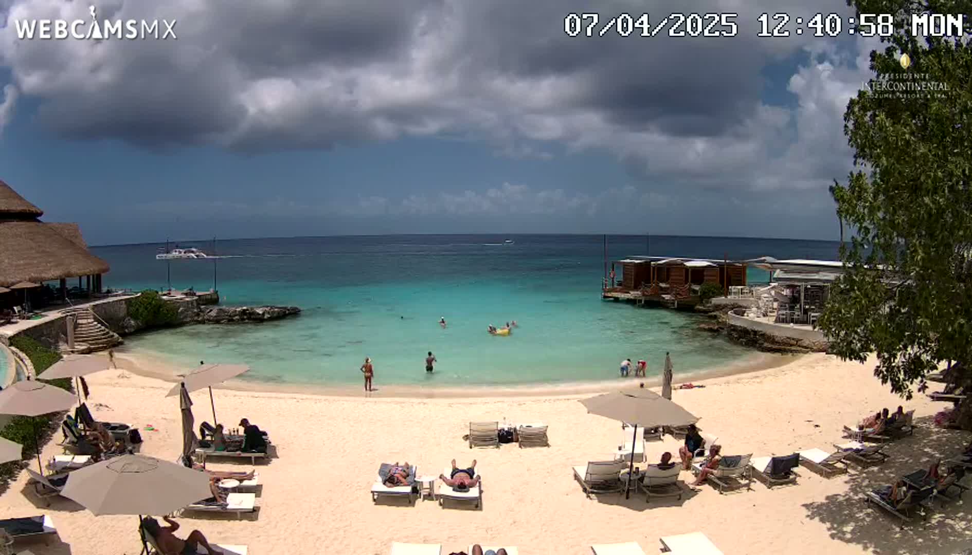 A partly cloudy day at a beach resort shows a sandy beach with lounge chairs and umbrellas, turquoise water, and several people swimming and sunbathing.