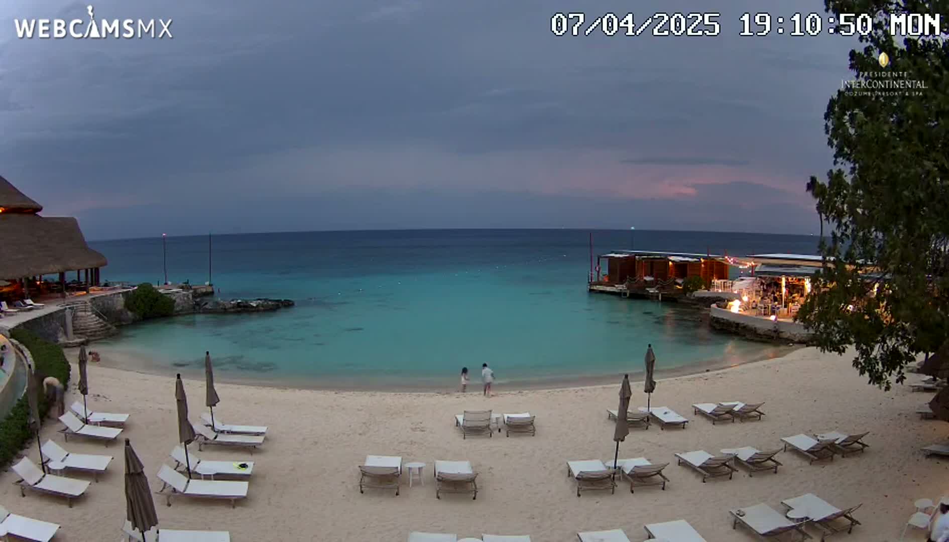 A tranquil beach scene at dusk shows a sandy shore lined with lounge chairs, two people wading in calm, turquoise water near a beachfront restaurant, and a thatched-roof building in the background under a cloudy sky.