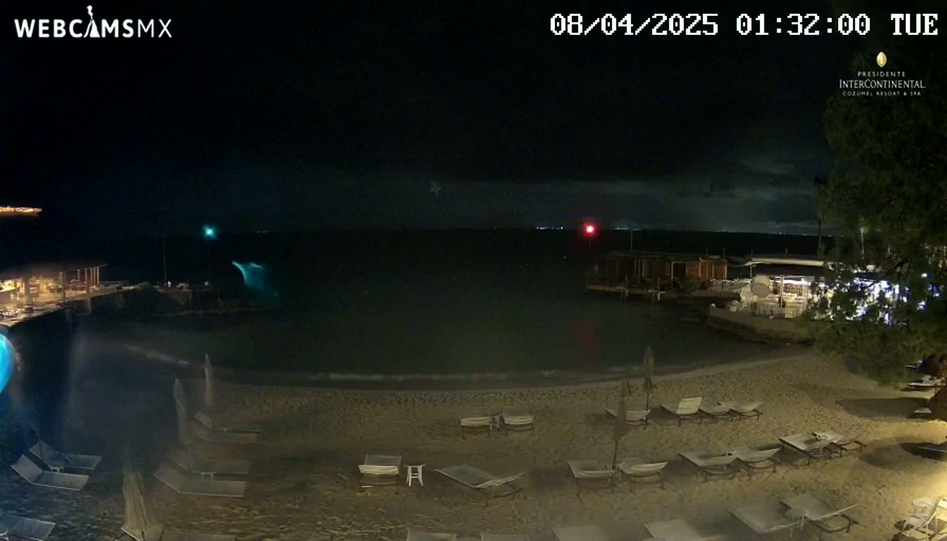 A nighttime beach scene shows empty lounge chairs on a sandy shore, with a dark ocean in the background under a mostly clear sky.