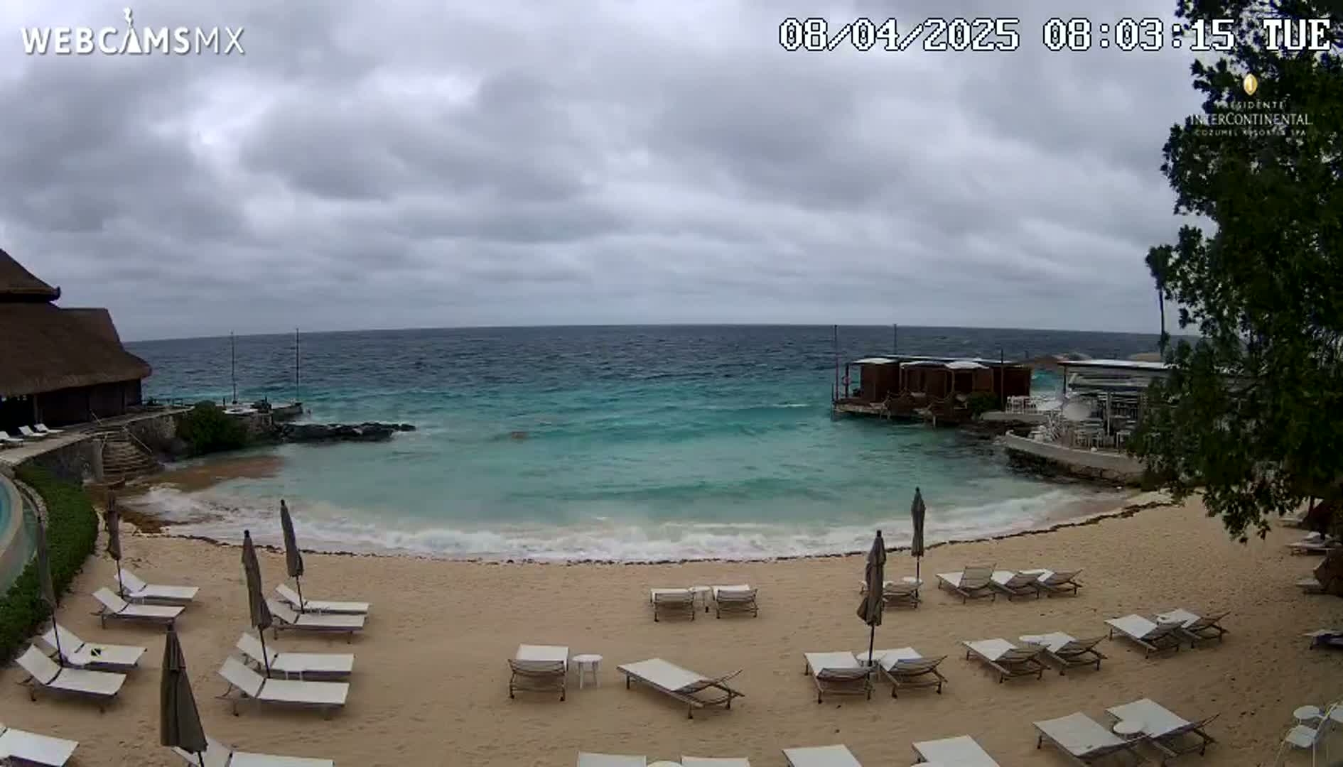 A sandy beach with lounge chairs and umbrellas is situated next to a calm, turquoise ocean under an overcast sky.