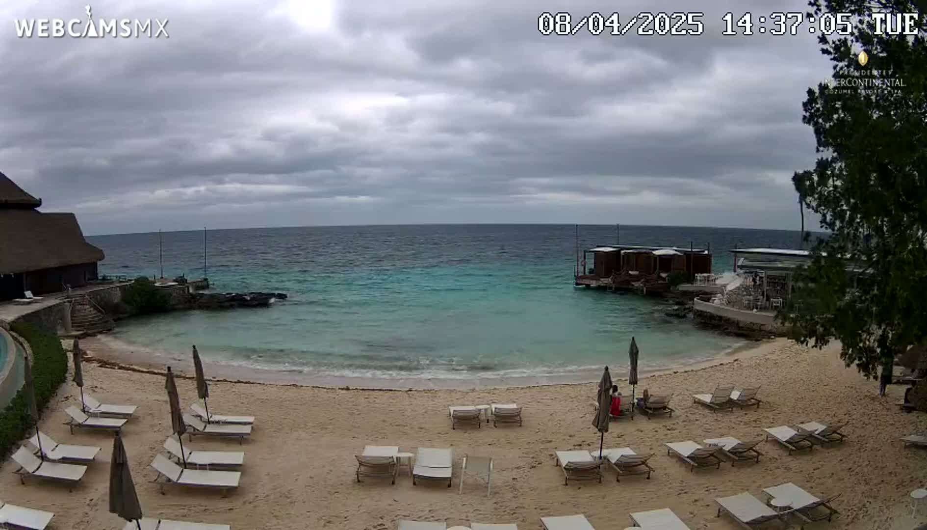 A sandy beach with lounge chairs and umbrellas overlooks calm, turquoise water under an overcast sky.