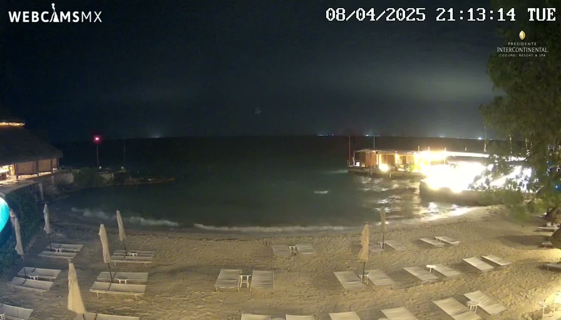 A nighttime beach scene shows calm waves lapping a sandy shore lined with empty lounge chairs and umbrellas, with a brightly lit structure visible across the water.