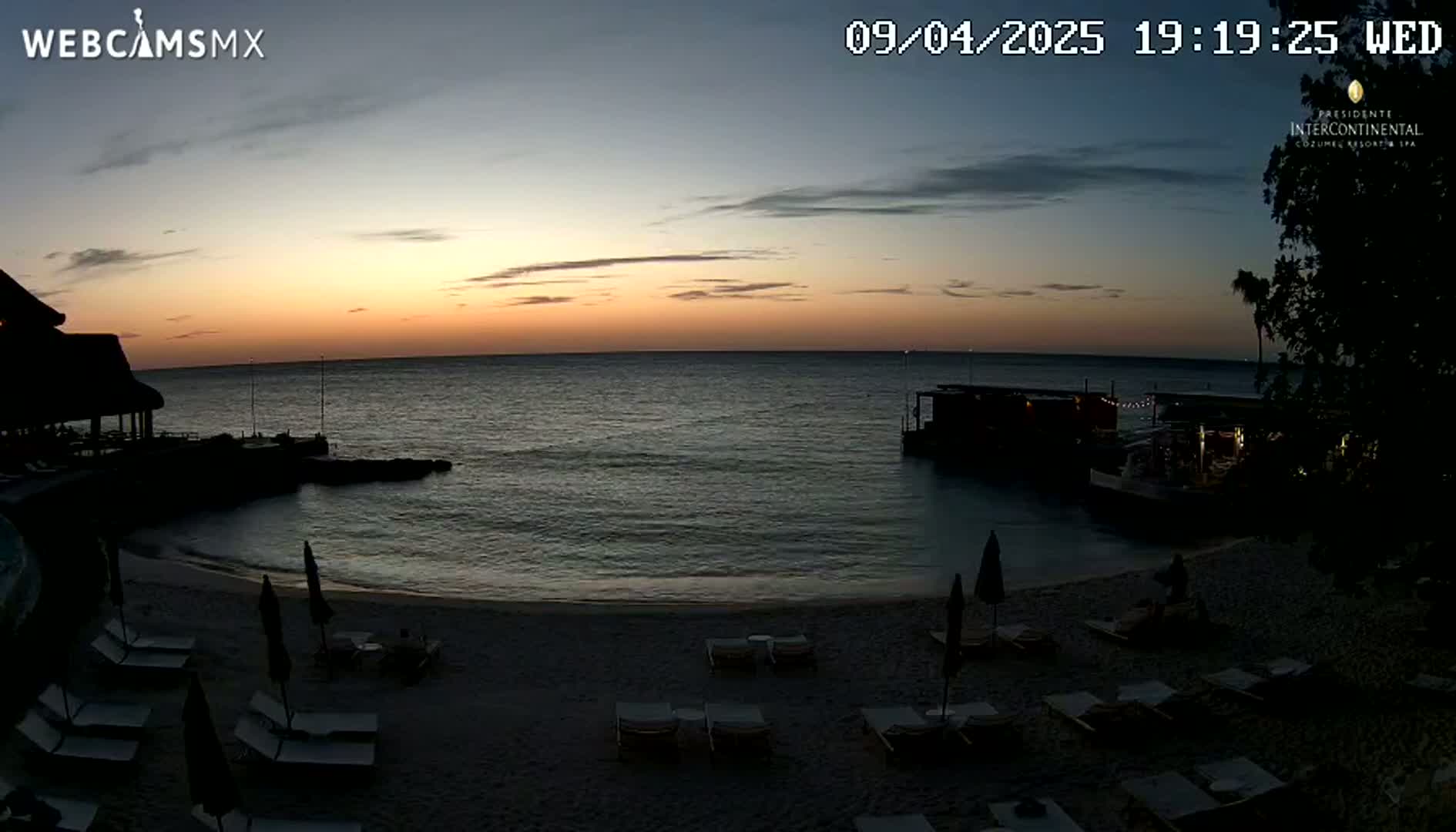 A beach scene at sunset shows calm water, a sandy shore with lounge chairs, and a structure resembling a bar or restaurant on the water's edge.