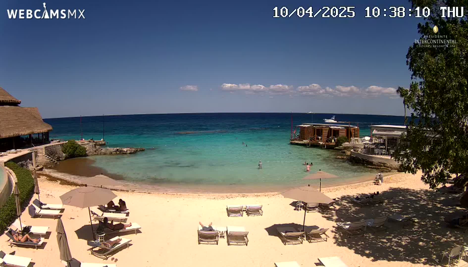 A sandy beach with lounge chairs and umbrellas under a mostly sunny sky,  borders a calm, turquoise ocean where people are swimming near a small overwater structure.