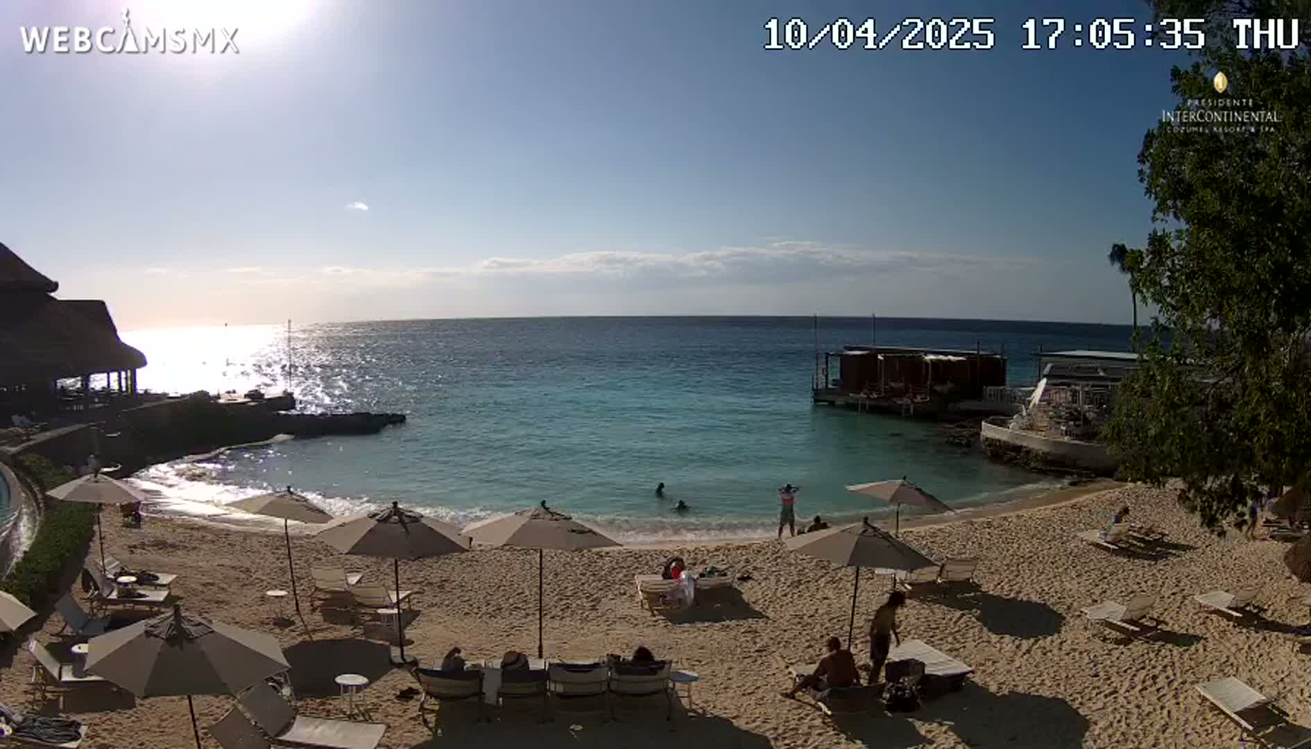 A sandy beach with lounge chairs and umbrellas under a sunny sky overlooks a calm, turquoise ocean, with a few people swimming and lounging.