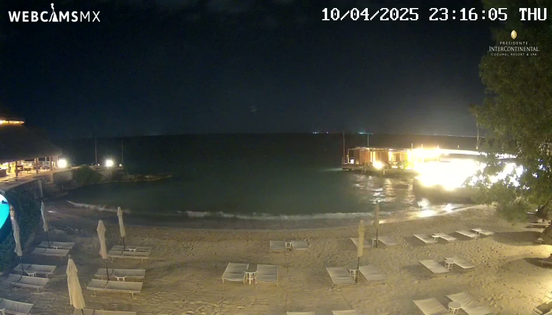 A nighttime beach scene shows gently lapping waves on a sandy shore with several empty lounge chairs under the dark sky.
