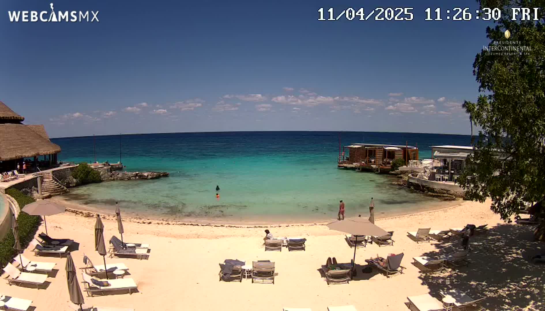 A sandy beach with lounge chairs and umbrellas sits next to calm, turquoise water, with a few people swimming and relaxing on the beach under a sunny sky.