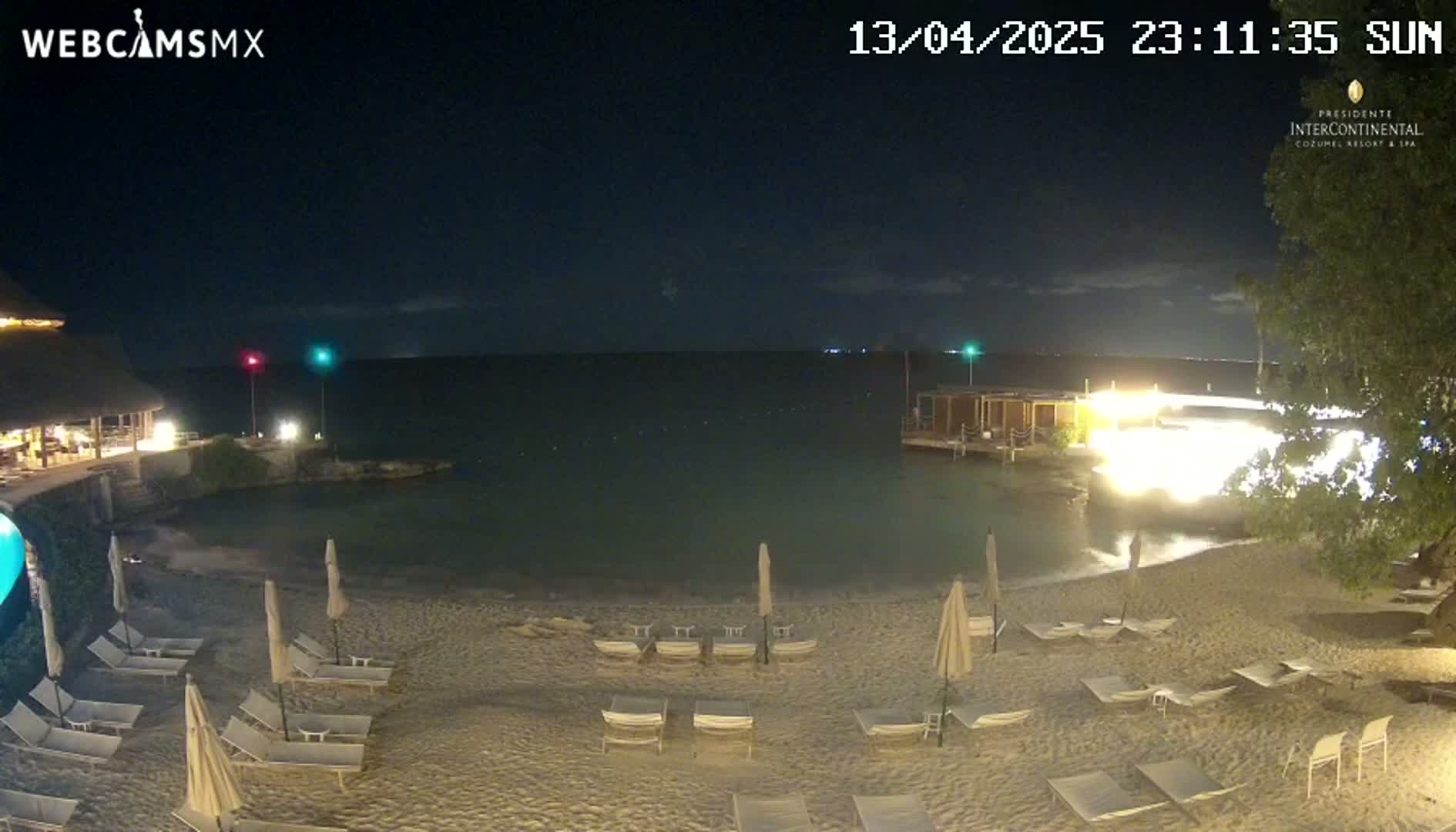 A nighttime beach scene shows a sandy shore with lounge chairs and umbrellas under a clear, dark sky, with calm water extending to a building with lights on in the distance.