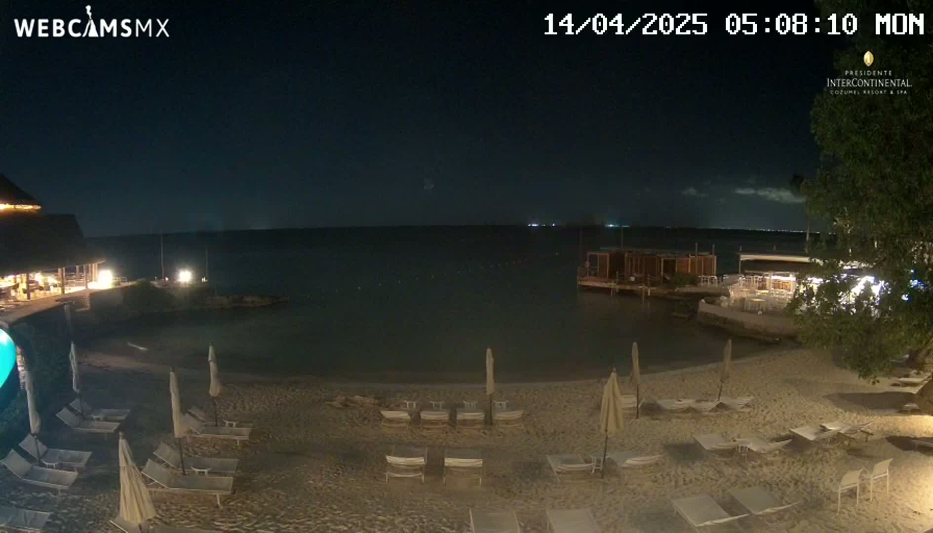A nighttime beach scene shows lounge chairs and umbrellas on a sandy beach next to calm water, with a building and vegetation visible in the background under a mostly clear sky.
