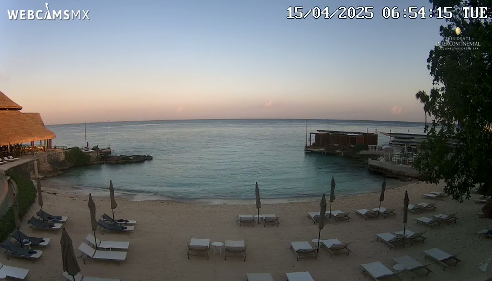 A tranquil beach scene at sunrise shows calm, turquoise water lapping a sandy shore lined with lounge chairs and umbrellas, a small structure on the water, and a thatched-roof building in the background.