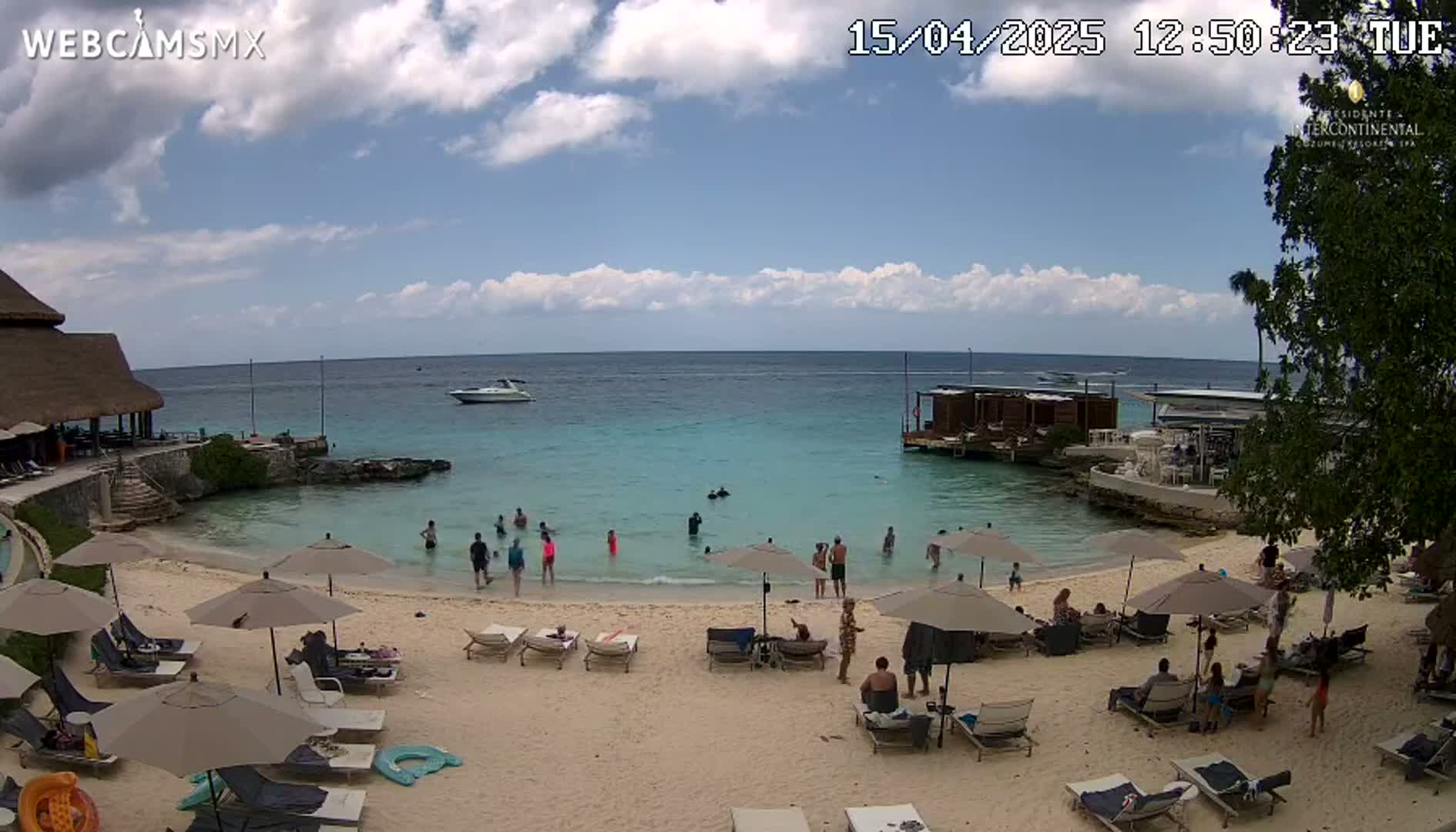 A sandy beach with lounge chairs and umbrellas, people swimming and relaxing in shallow turquoise water, a boat in the distance, and partly cloudy skies.