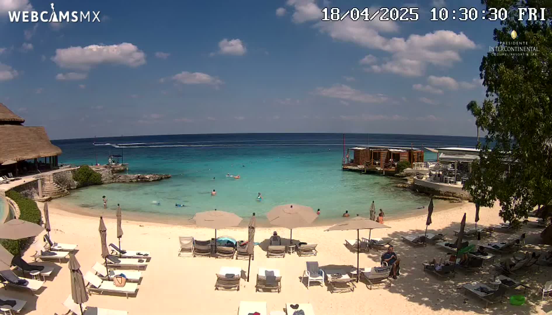 A beach scene with calm, turquoise water, a sandy shore dotted with lounge chairs and umbrellas, a few people swimming and lounging, and a thatched-roof structure in the background under a partly cloudy sky.