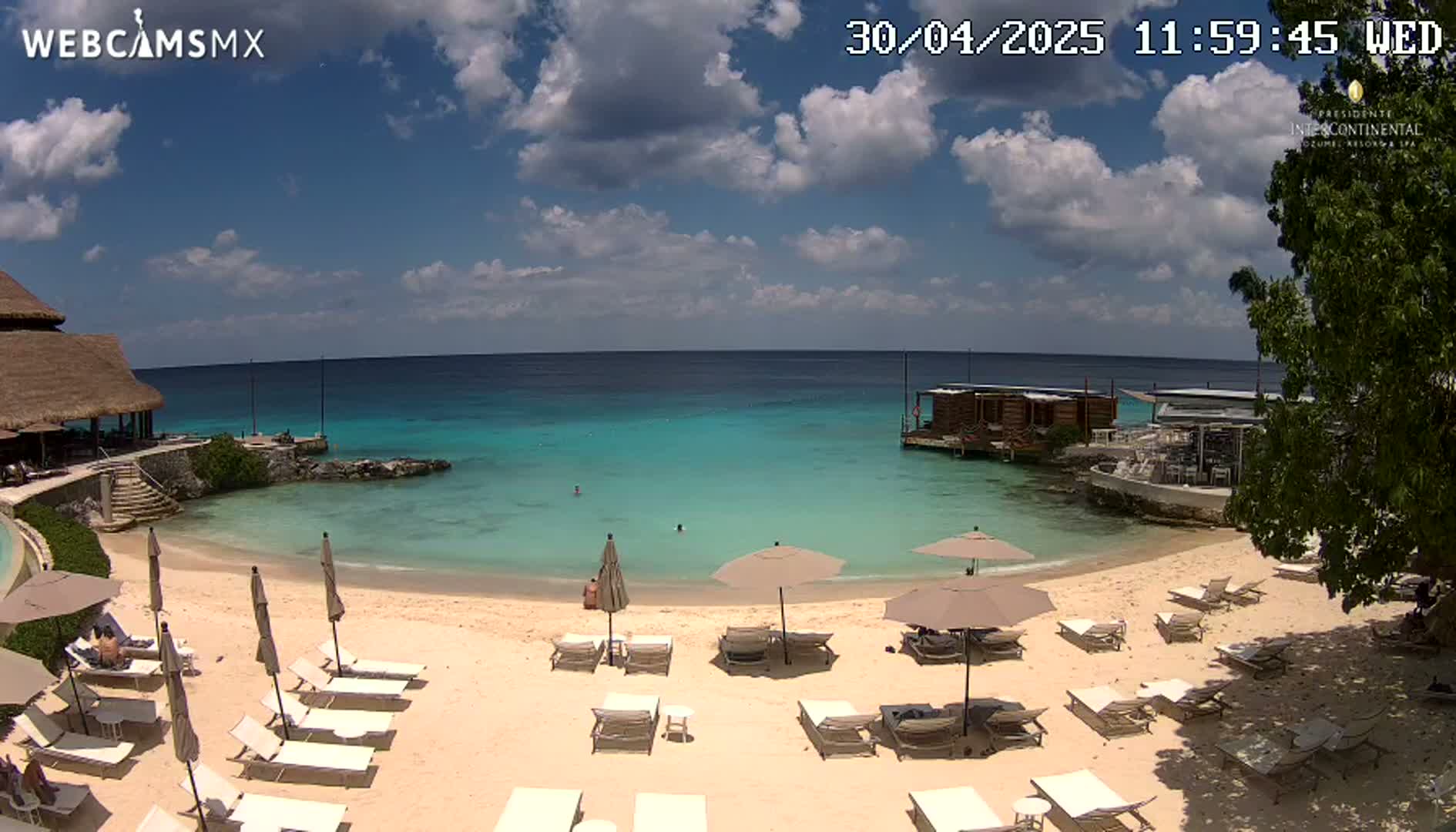 A partly cloudy sunny day shows a beach with lounge chairs and umbrellas, a calm turquoise ocean, and a beachfront building.