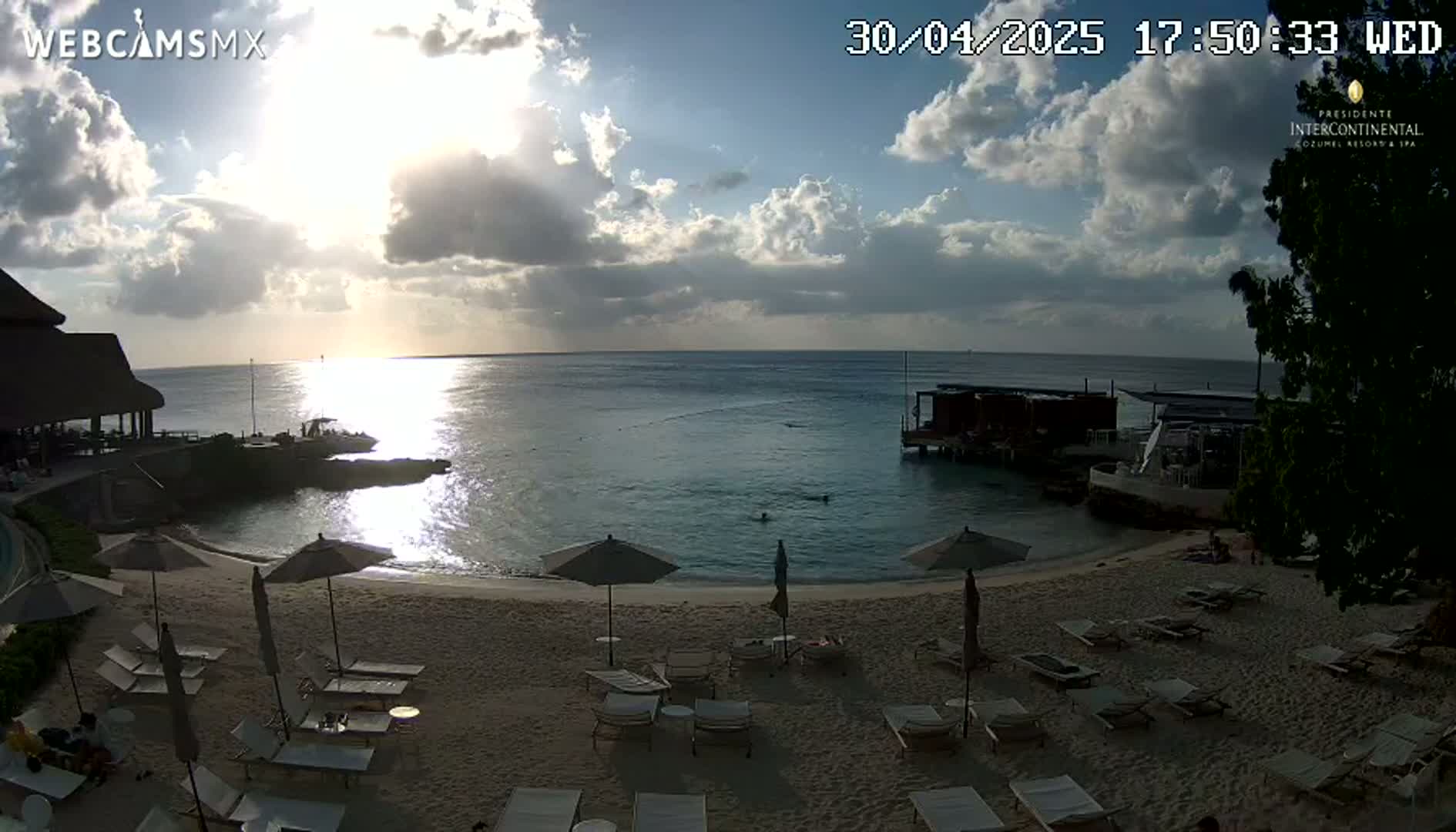 A partly cloudy sunset illuminates a beach with lounge chairs and umbrellas, overlooking calm ocean water and a resort structure.