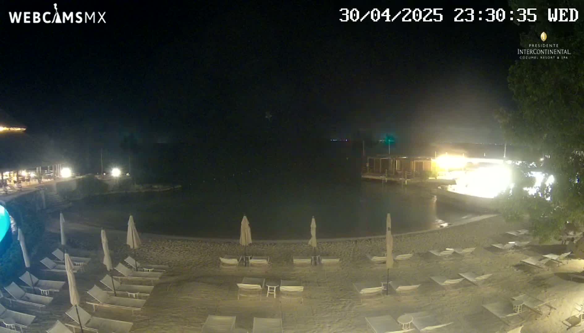 A nighttime view of a beach with lounge chairs and umbrellas surrounding a calm body of water, illuminated by lights from nearby buildings.