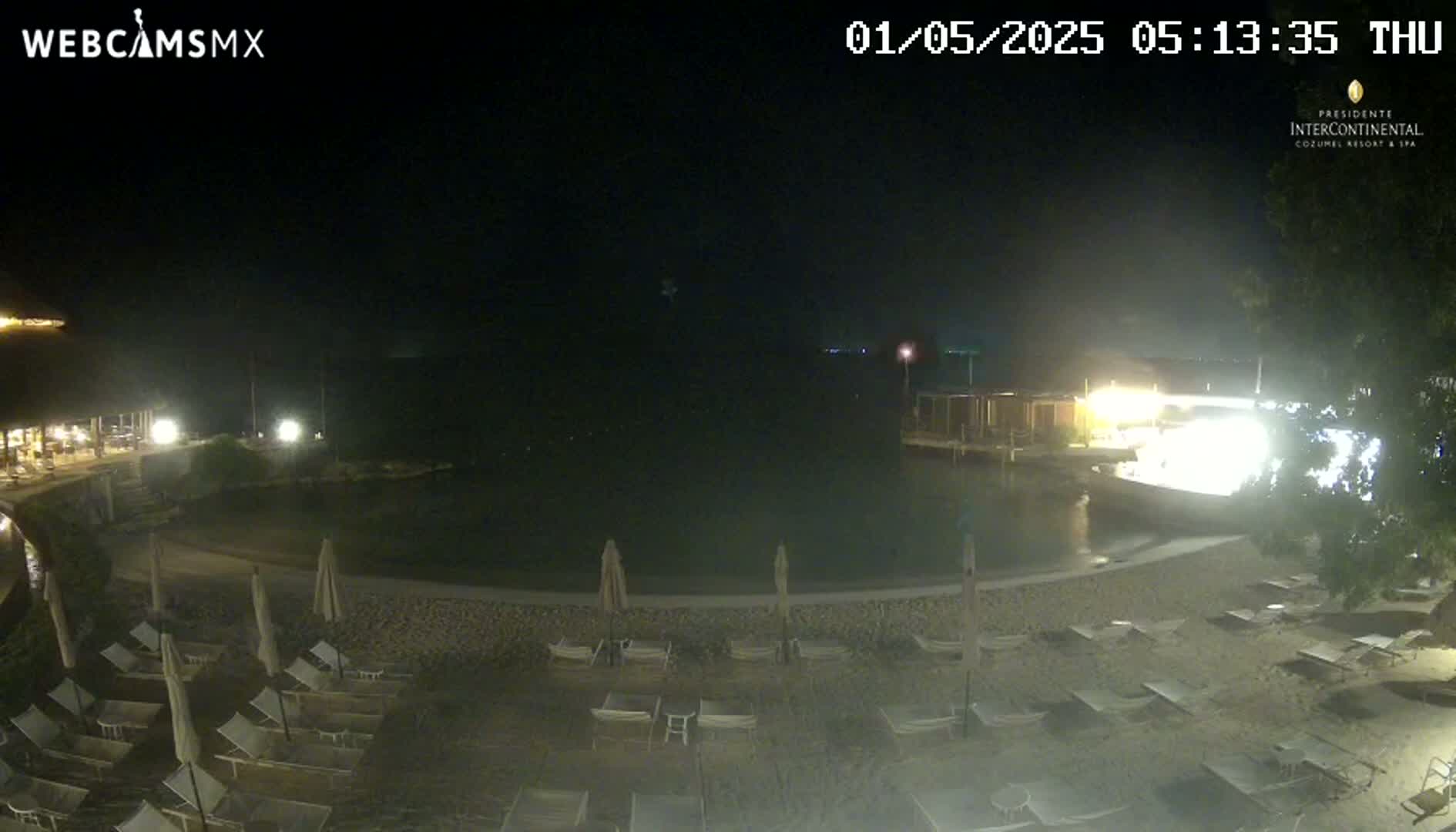 A nighttime view of a resort beach area with a calm body of water, numerous lounge chairs, and some buildings illuminated by lights.
