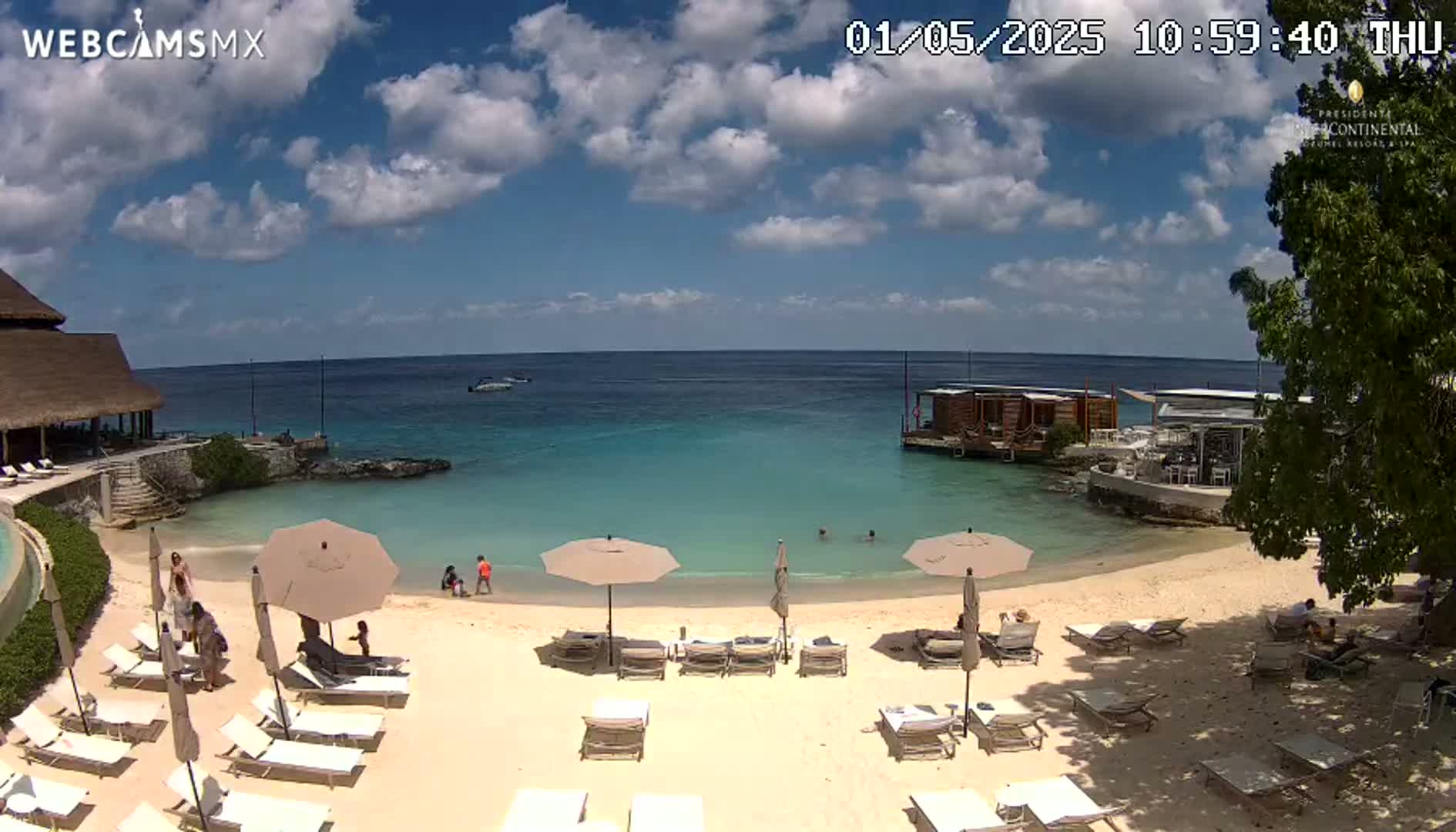 A partly sunny beach scene shows a sandy shore with lounge chairs and umbrellas, calm turquoise water, a small building over the water, and a few people enjoying the beach.
