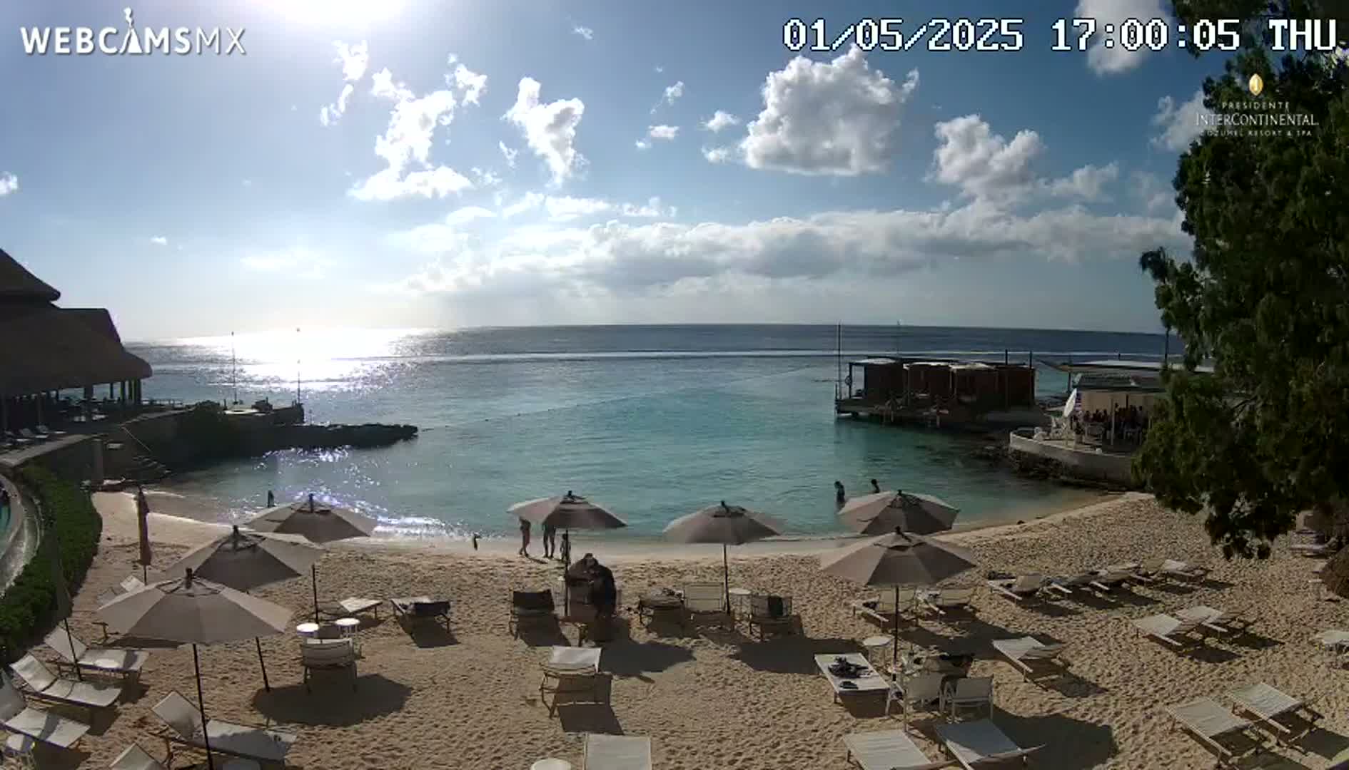 A sandy beach with lounge chairs and umbrellas under a partly sunny sky overlooks a calm, turquoise ocean.