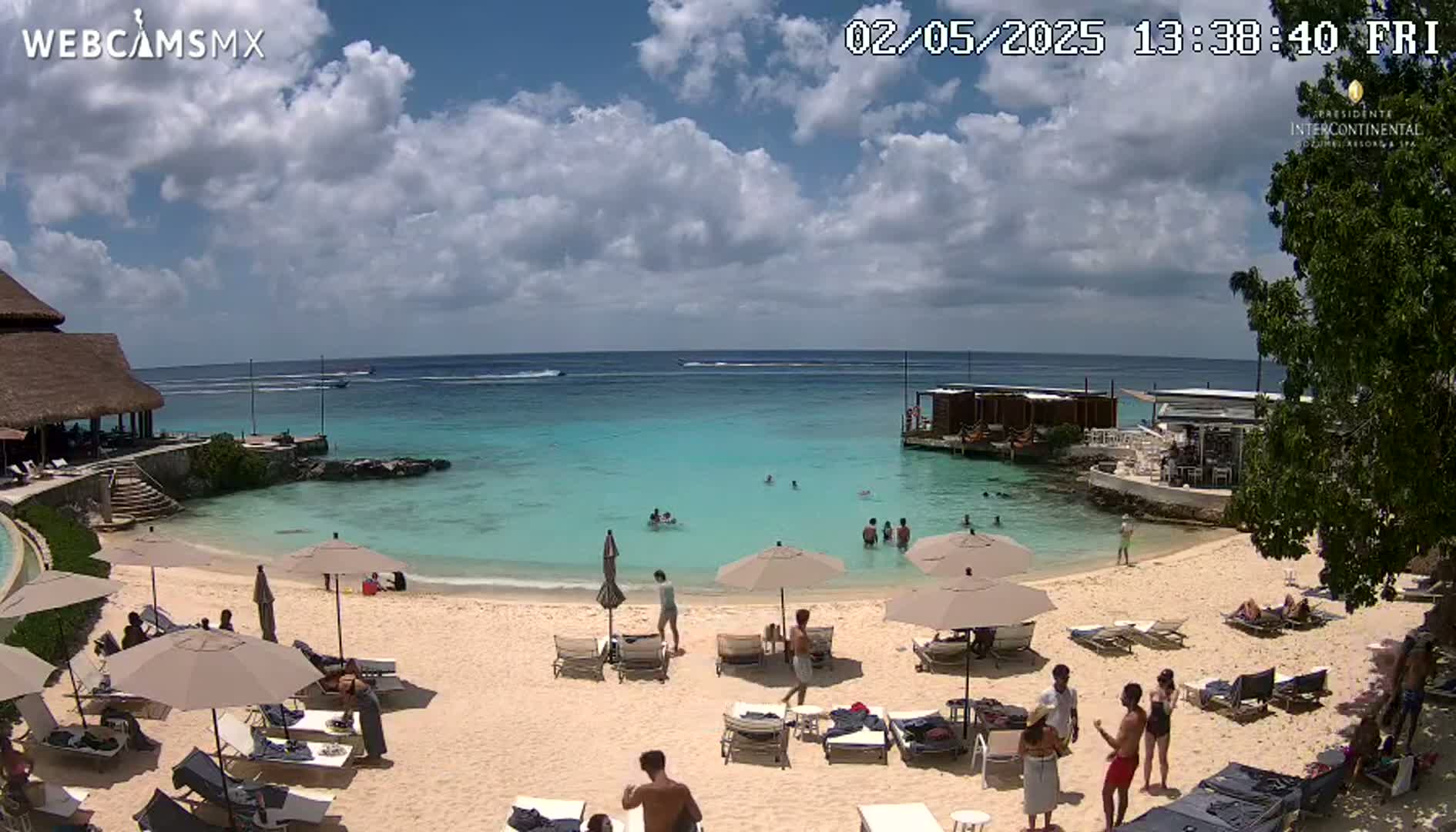 A partly cloudy sunny day shows a beach scene with turquoise water, people swimming and relaxing on lounge chairs under umbrellas on a sandy beach near a resort.