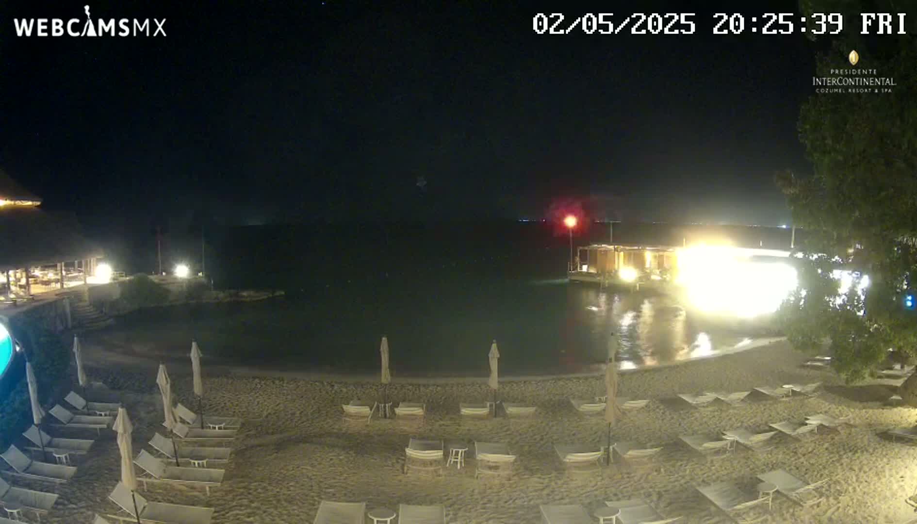 A nighttime view of a sandy beach with lounge chairs and umbrellas, a calm body of water, and buildings with lights visible in the distance.
