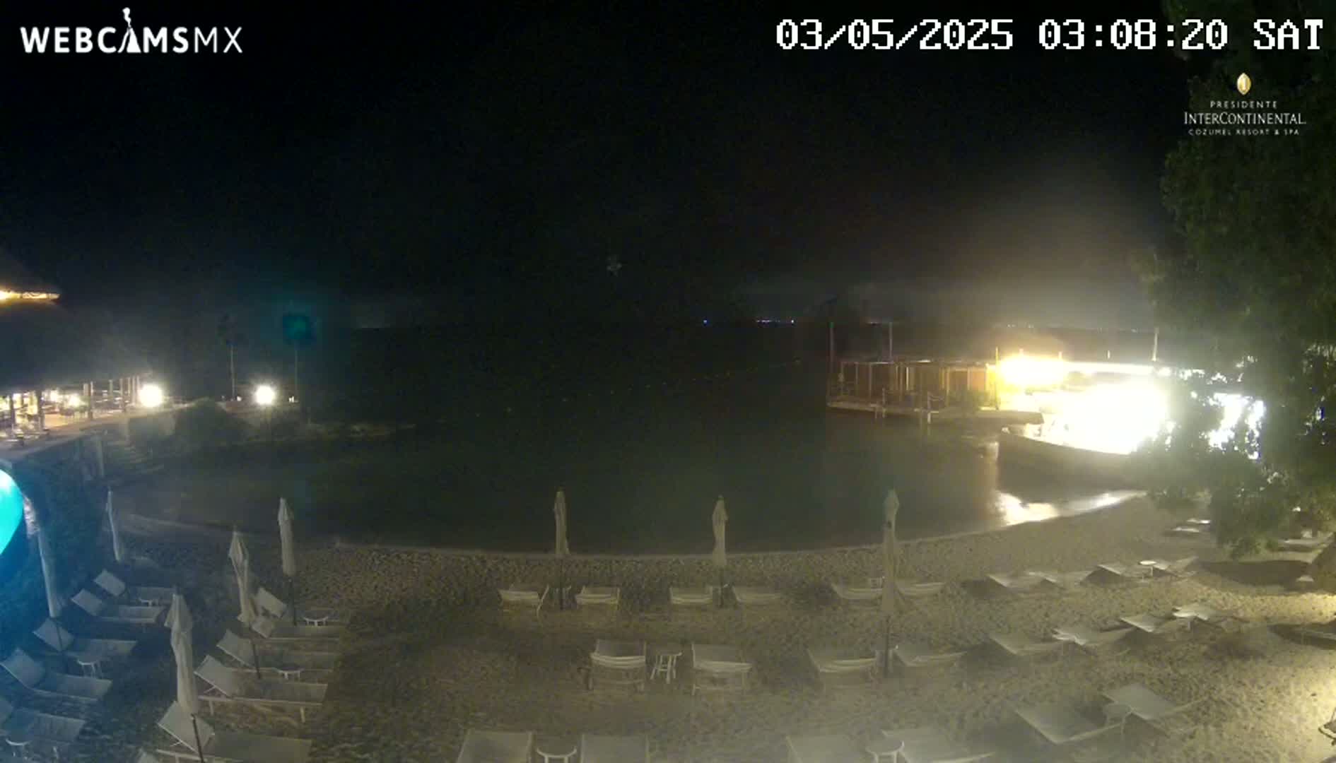 A nighttime view of a sandy beach with lounge chairs and umbrellas surrounding a calm body of water, illuminated by artificial lights.