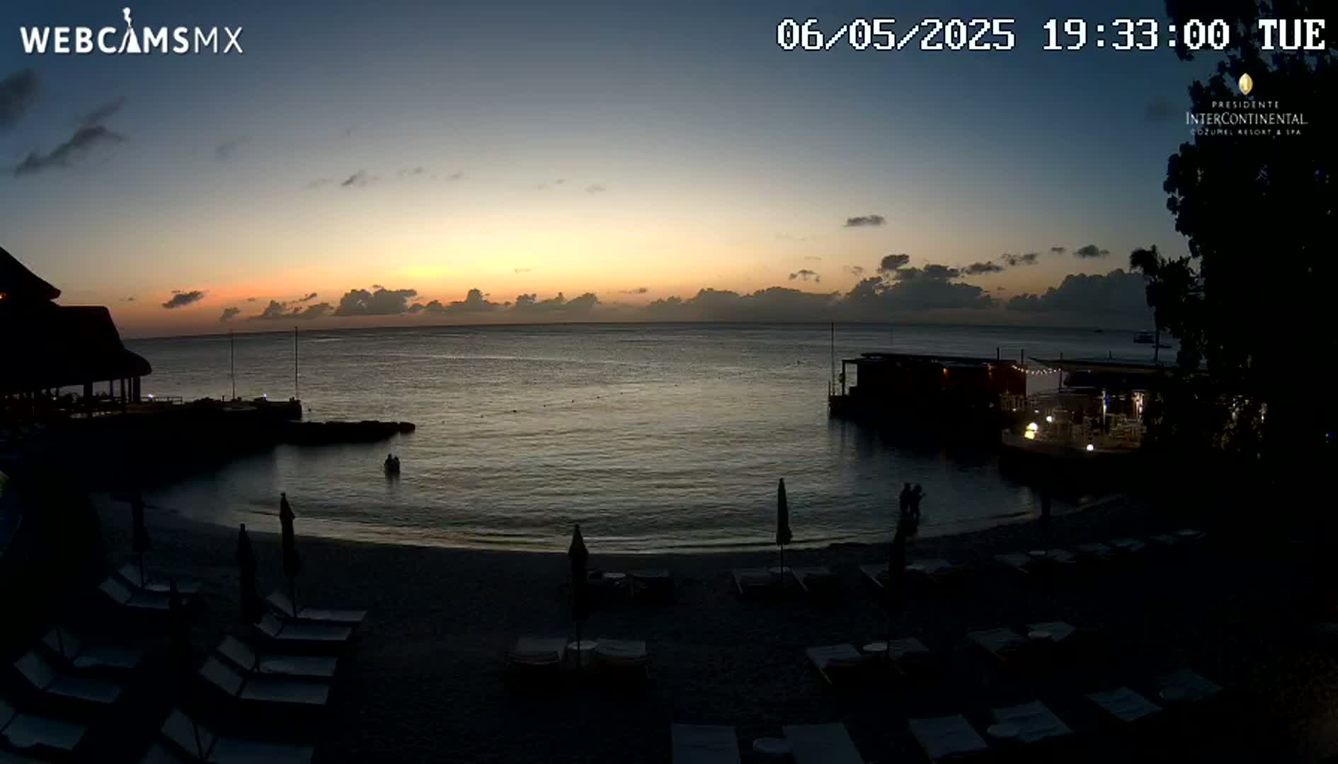 A beach at sunset with calm water, a few people, and lounge chairs.