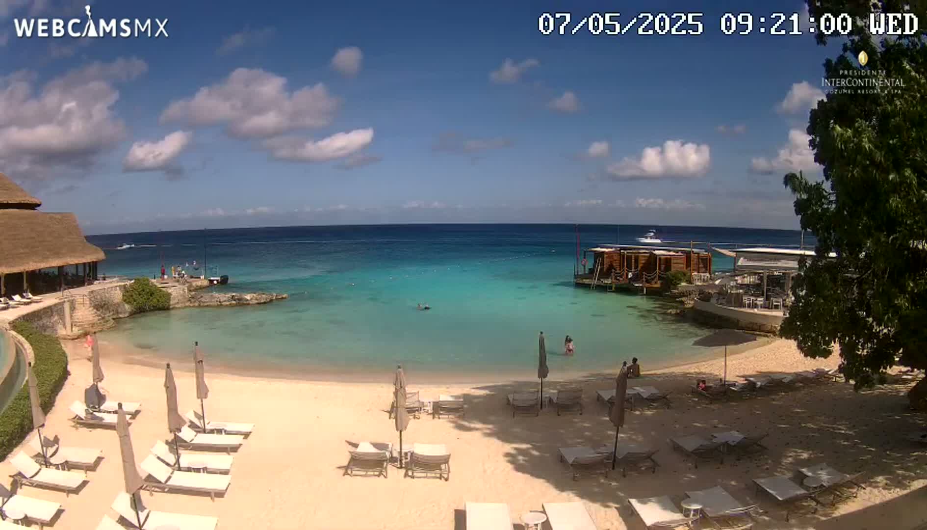 A partly sunny beach scene shows calm, turquoise water, a sandy beach with lounge chairs and umbrellas, a small overwater structure, and a thatch-roofed building.