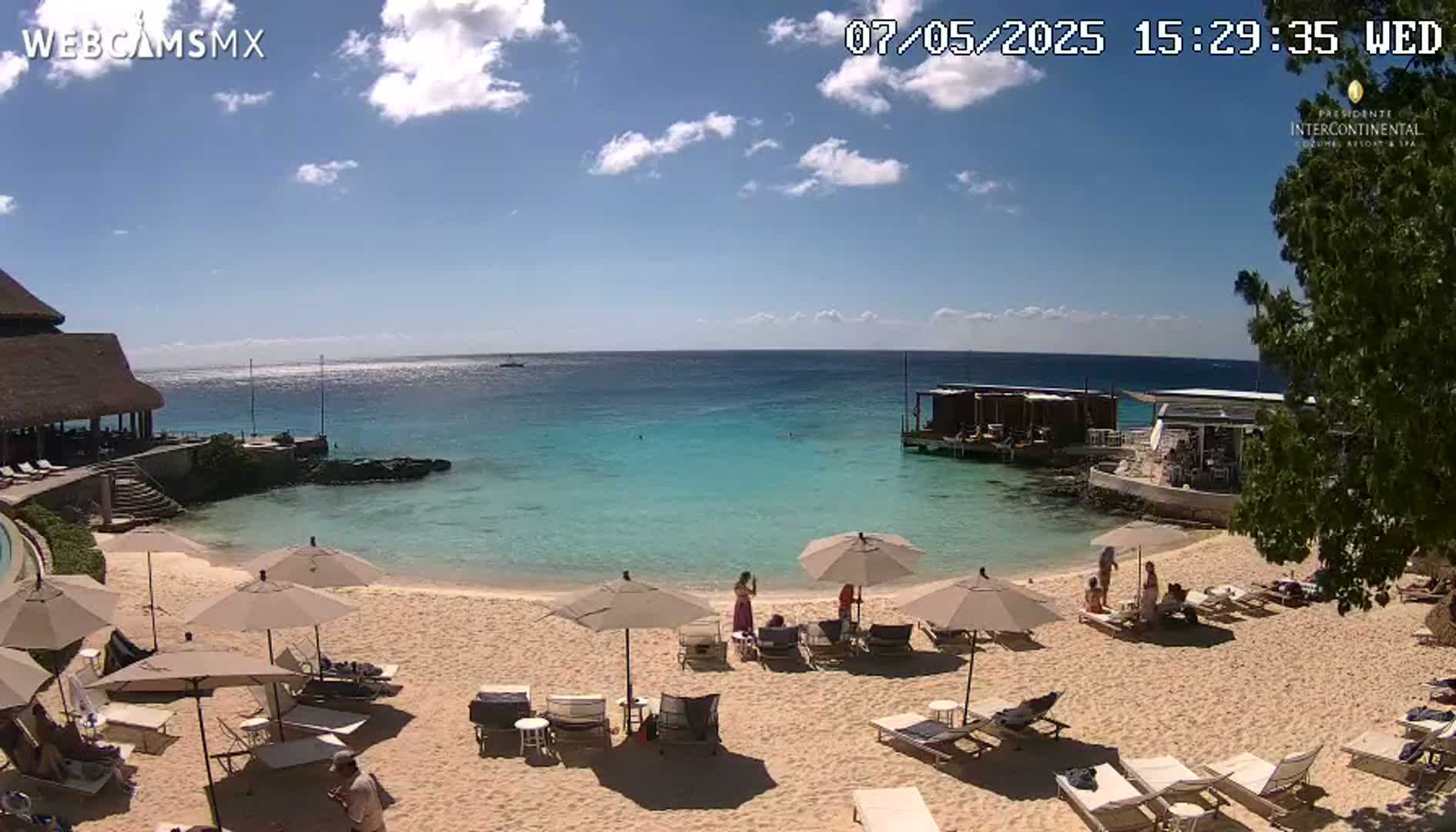 A sandy beach with turquoise water, beach umbrellas, and lounge chairs is seen on a sunny day with a few scattered clouds.